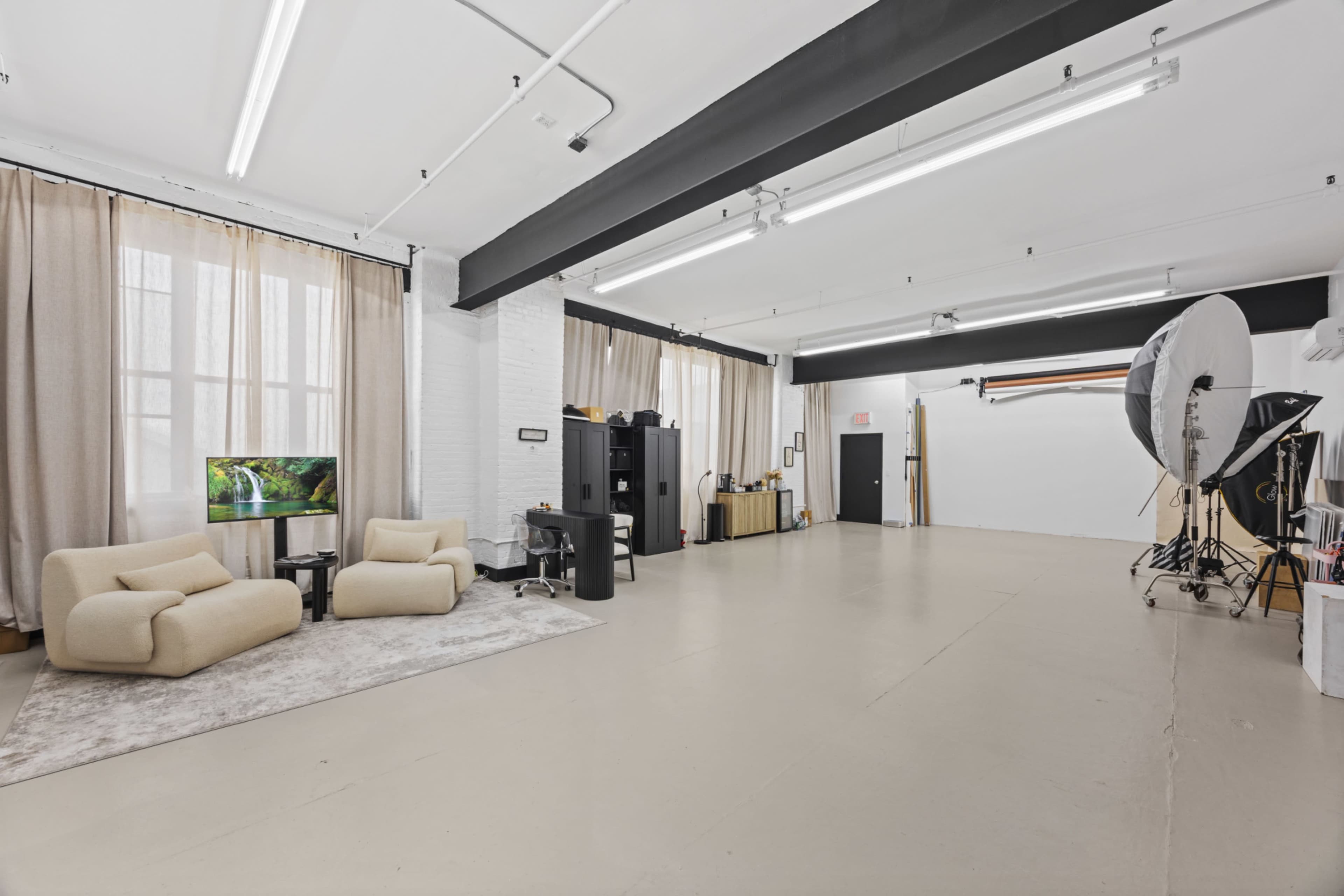 The image shows a spacious photography studio with soft beige furniture, large windows covered by drapes, and various photography equipment positioned in the corner.