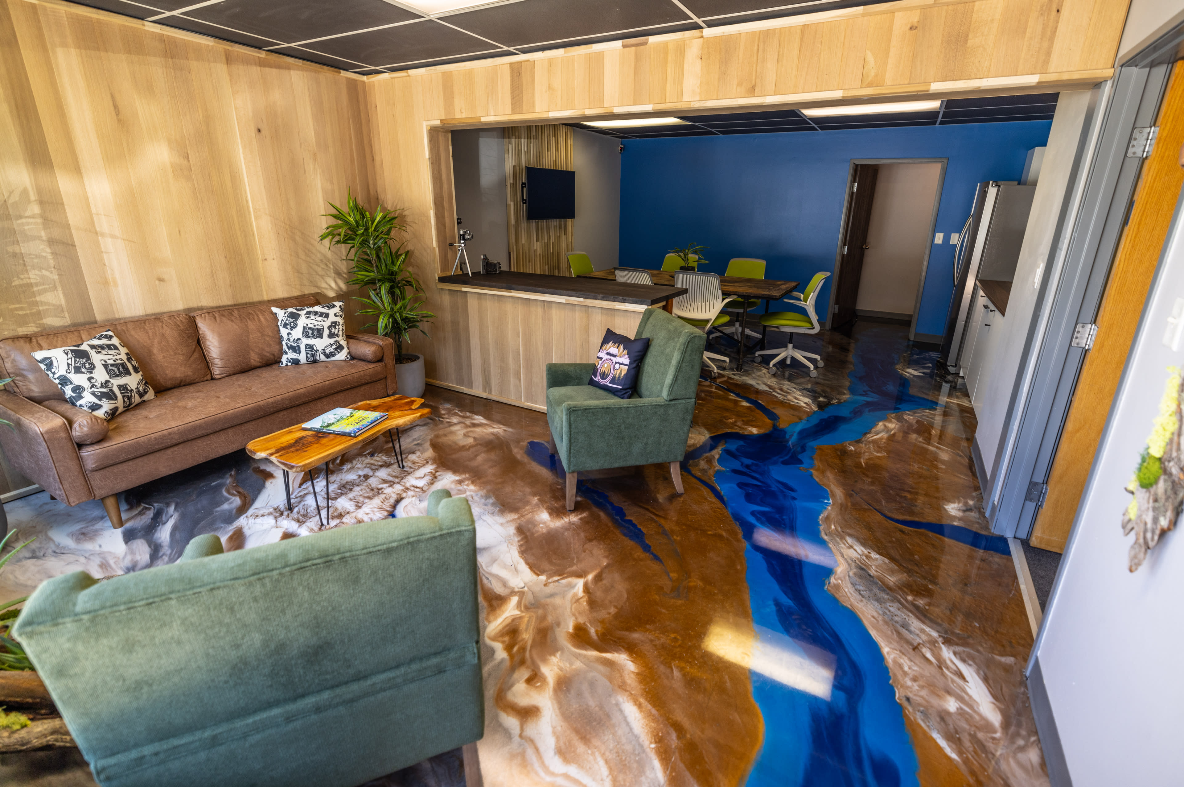 The image shows a modern office space featuring a cozy seating area with a brown sofa and green armchairs, alongside a kitchen and collaborative workspace with bright blue walls and vibrant furniture.