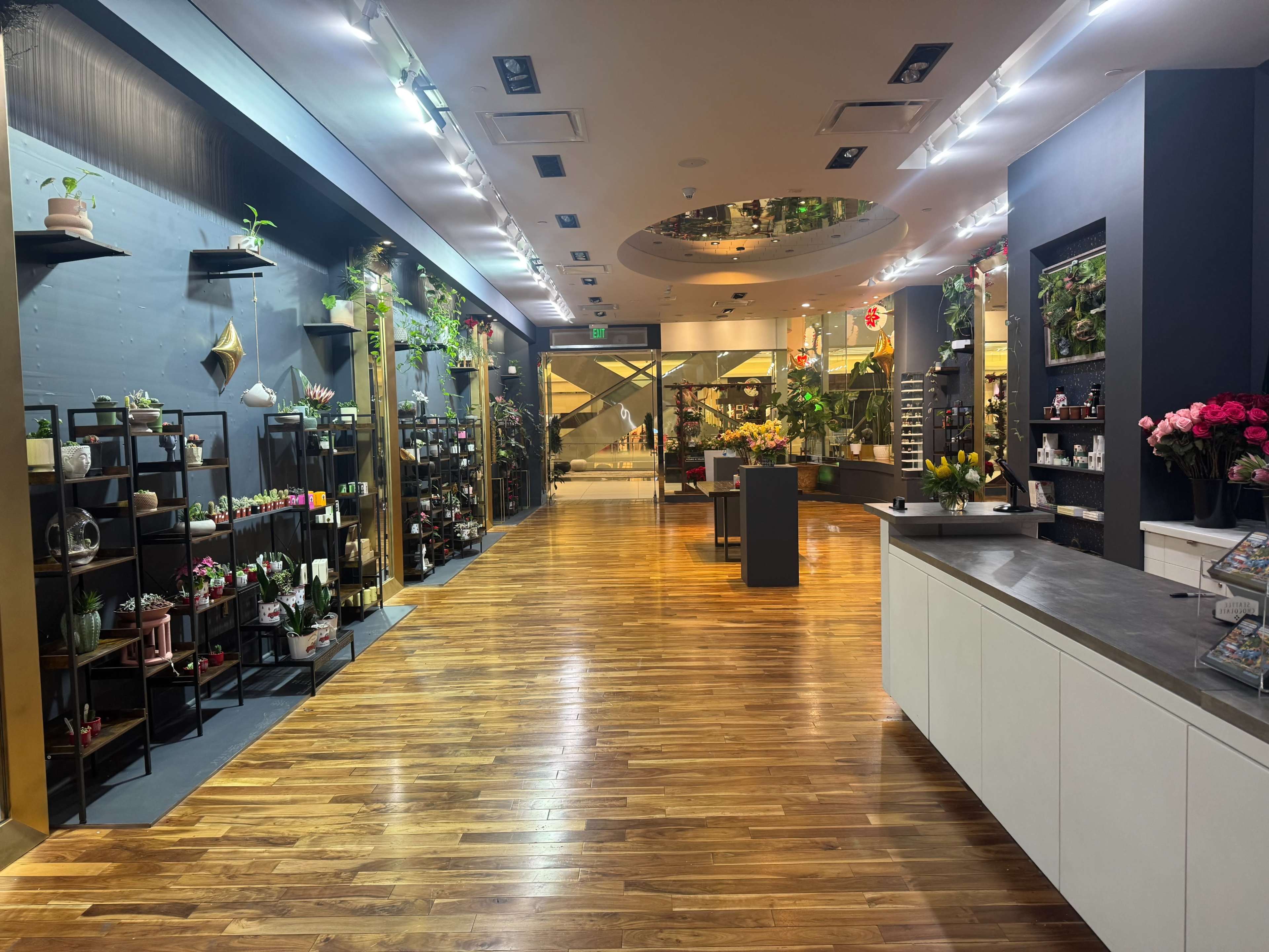 The image shows the interior of a floral shop with wooden flooring, rows of shelves filled with plants and floral arrangements, and a checkout counter.