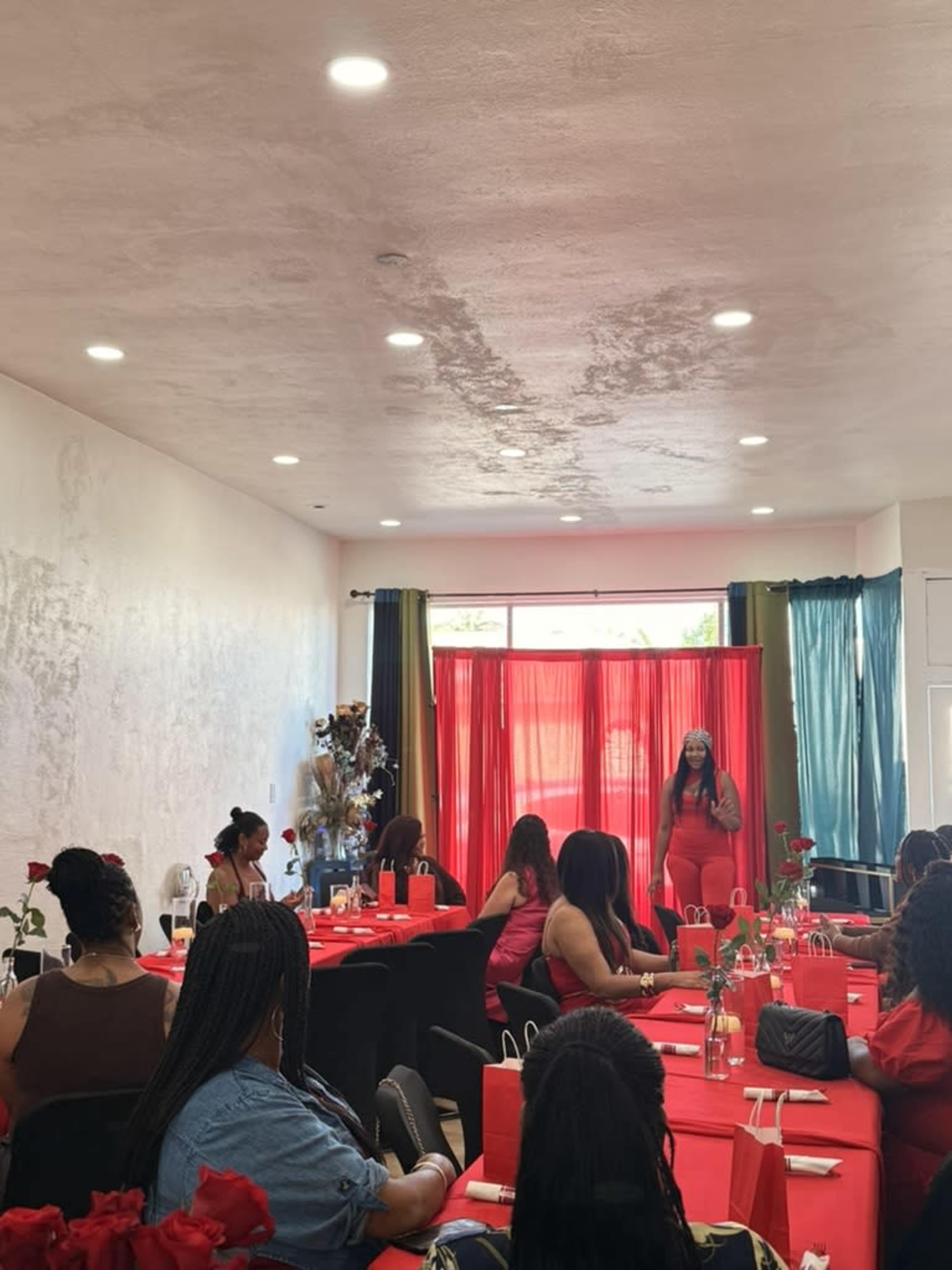 A woman in a red outfit speaks at the front of a room decorated with red tablecloths and floral centerpieces, while attendees listen attentively from seated positions.
