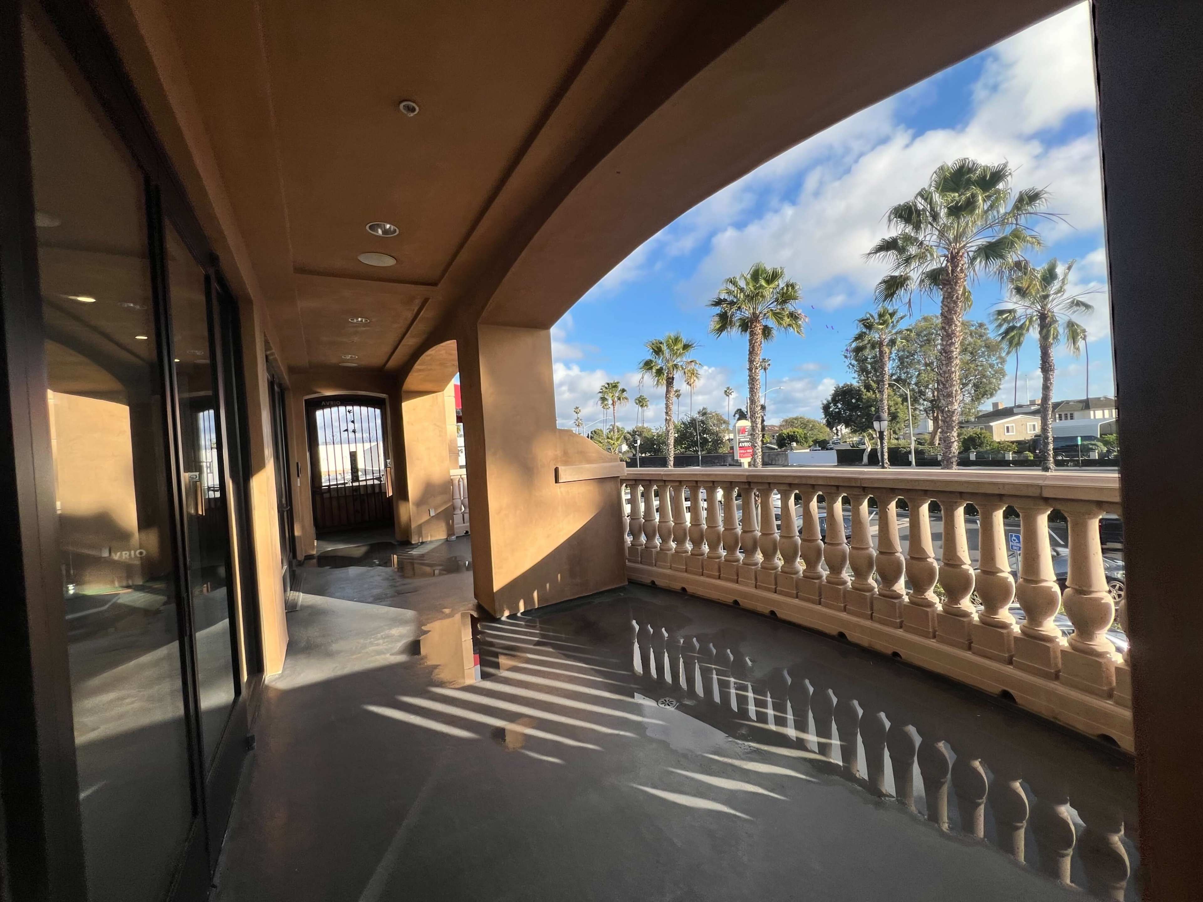 A covered balcony with concrete flooring and palm trees visible in the background under a partly cloudy sky.