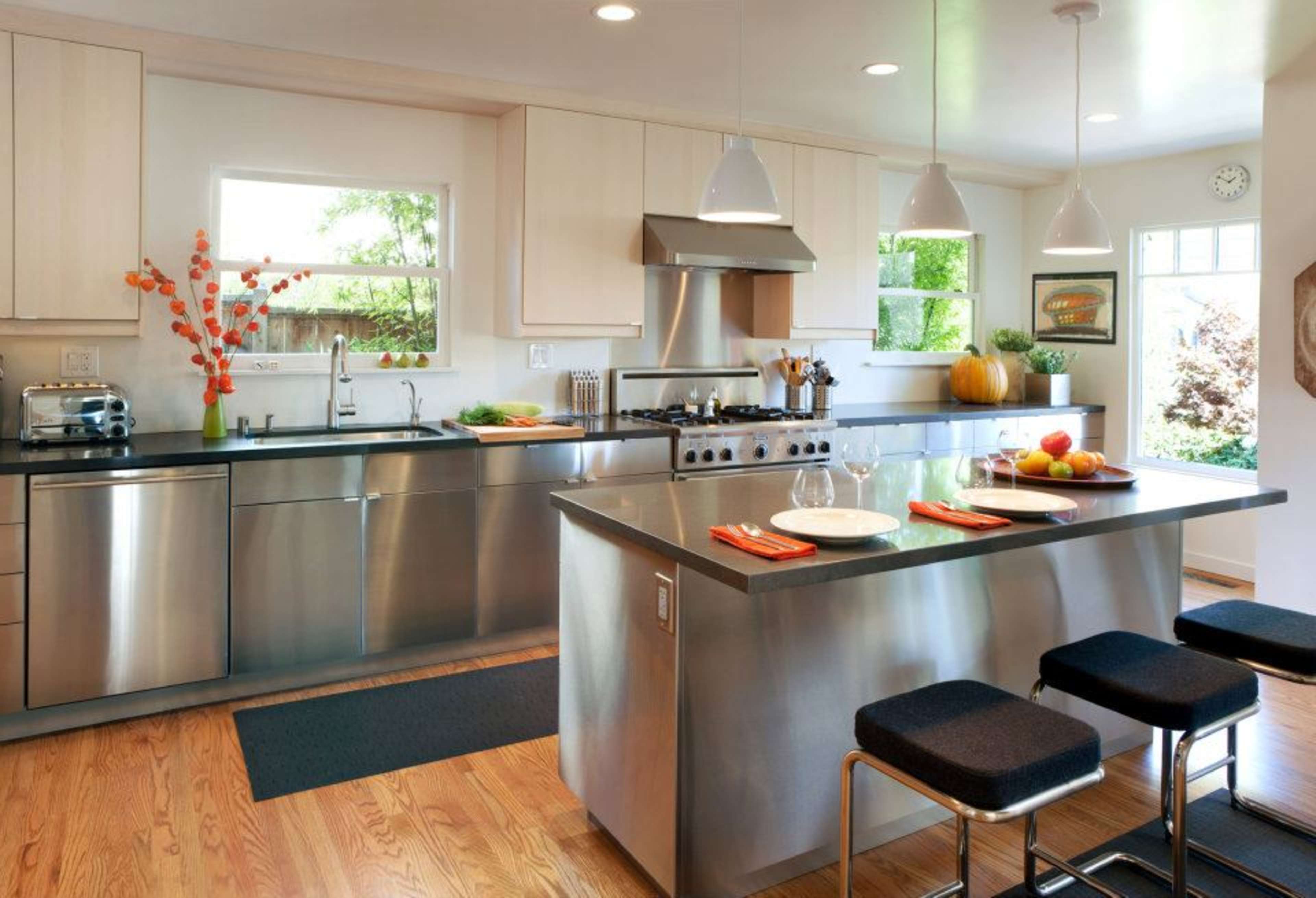 The kitchen features stainless steel cabinets and appliances, a central island with black stools, and large windows that provide natural light.