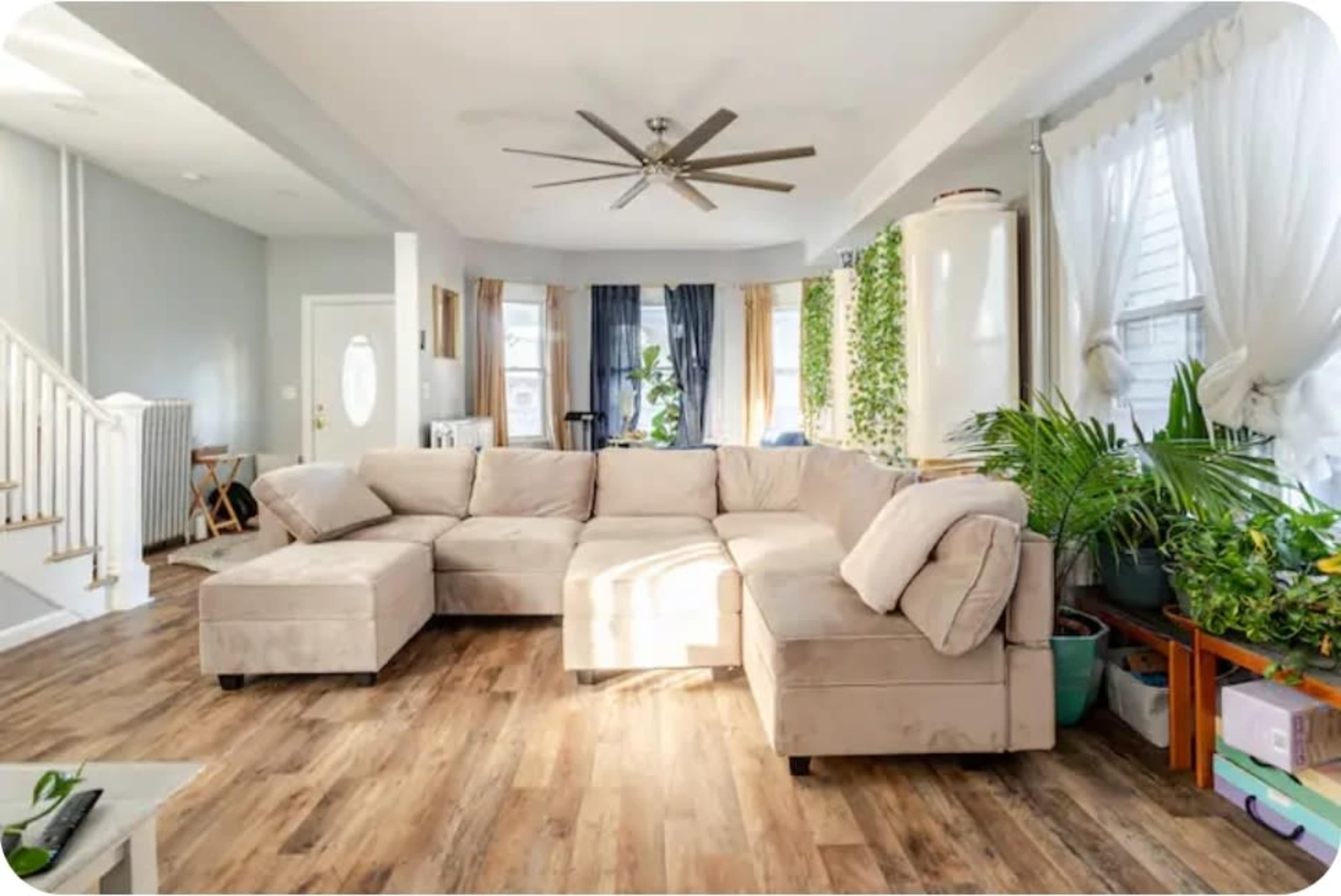 The image shows a spacious living room featuring a large beige sectional couch, wooden flooring, and several potted plants near a window.