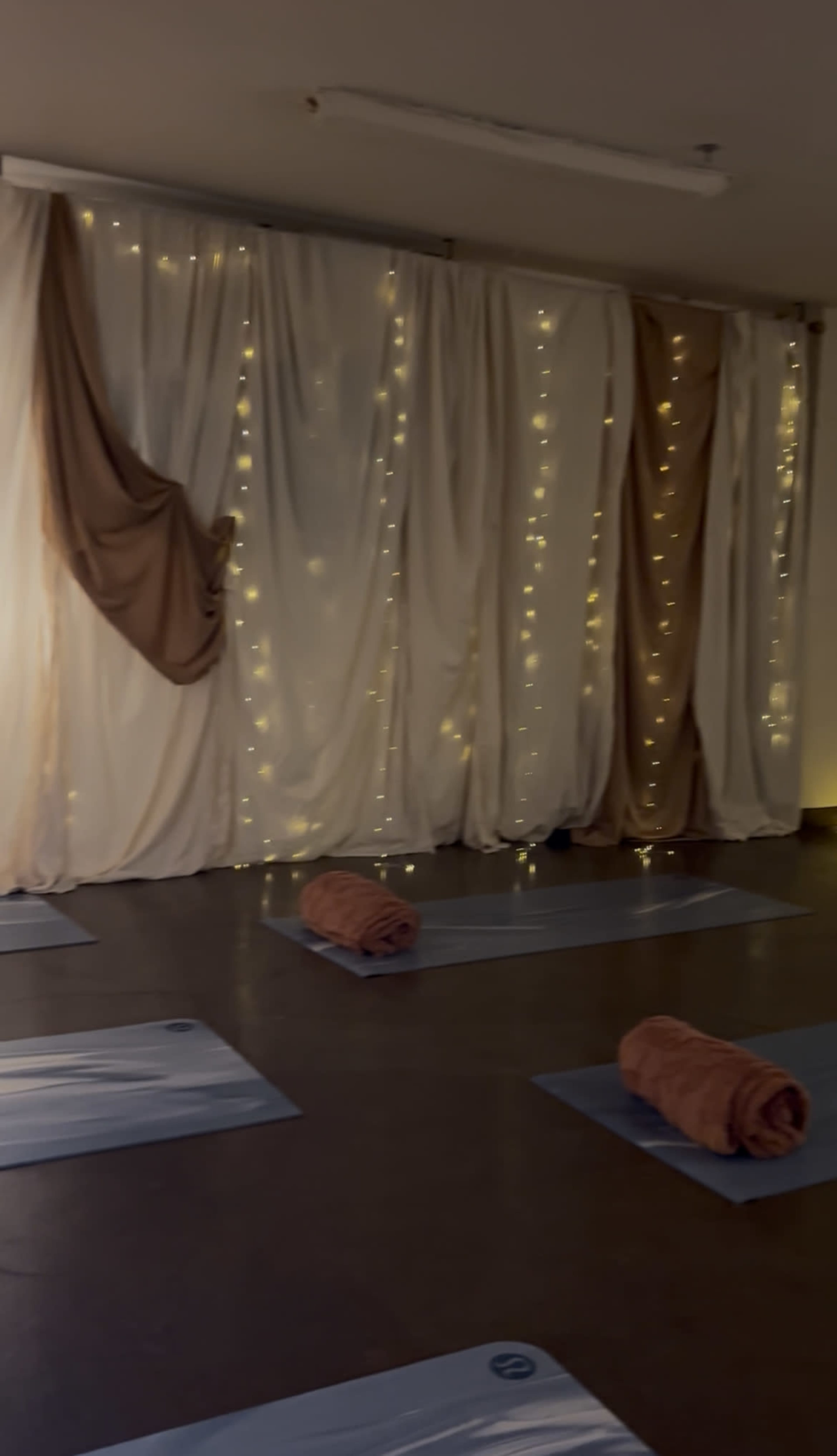 The image shows a serene yoga studio with rolled towels placed on exercise mats and curtains draped with twinkling fairy lights.