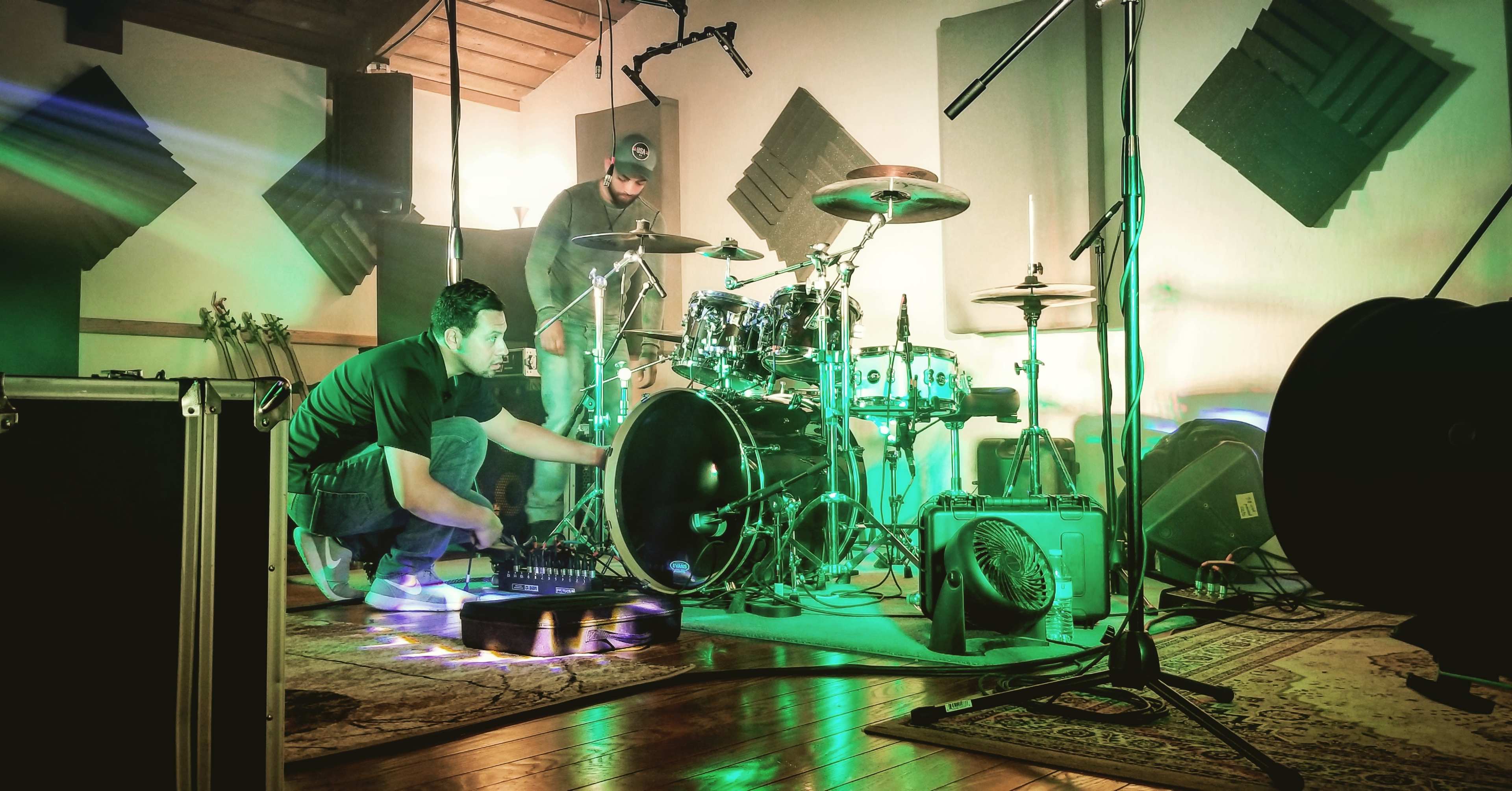Two musicians set up equipment in a recording studio with drums and microphones arranged in a well-lit space.