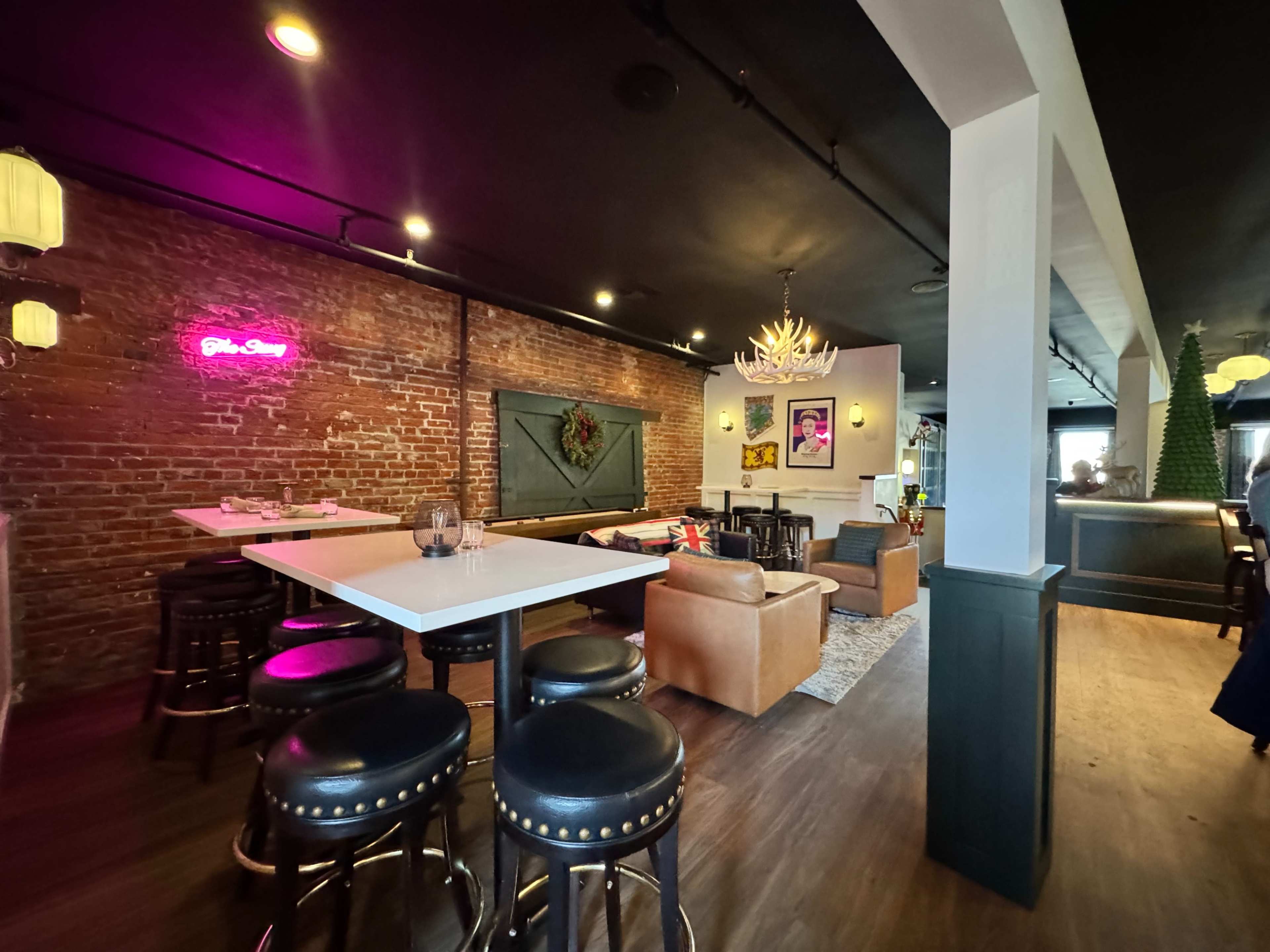 The image shows a modern lounge area featuring exposed brick walls, white tables with black stools, and a mix of casual seating, illuminated by decorative lighting.