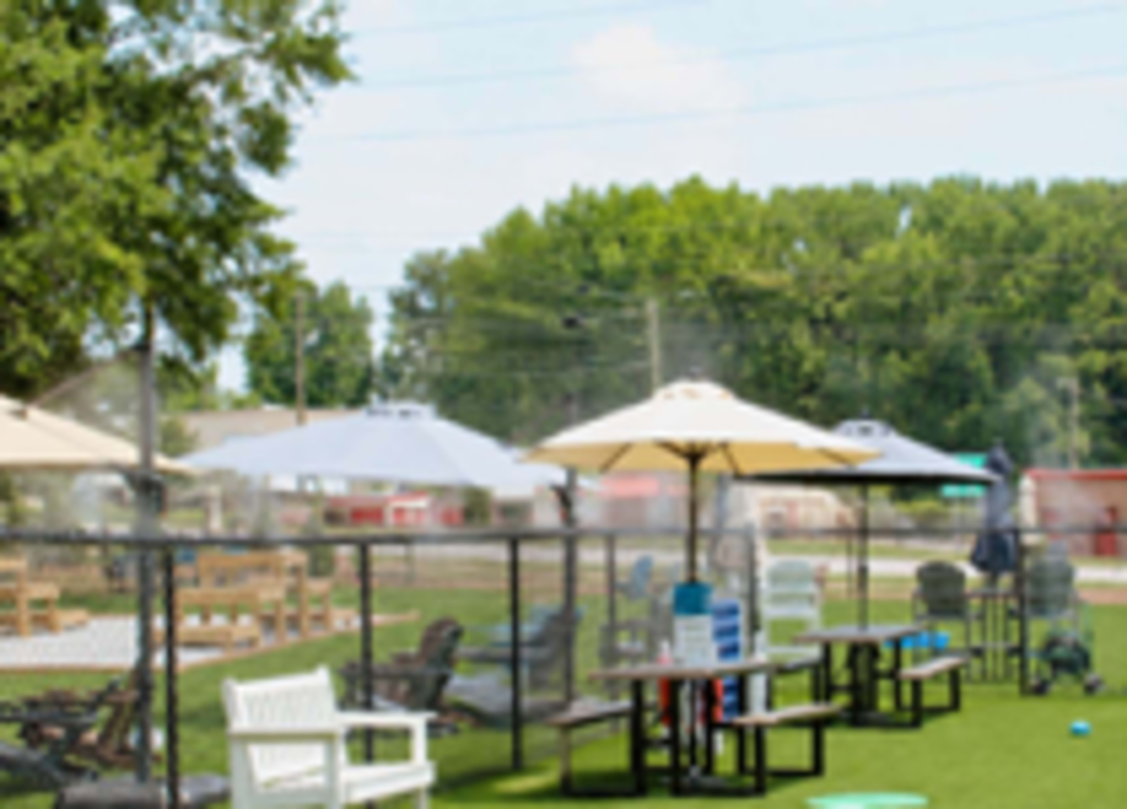 The image shows an outdoor area with tables, chairs, and umbrellas set up on a grassy surface, surrounded by a fence and misting equipment.