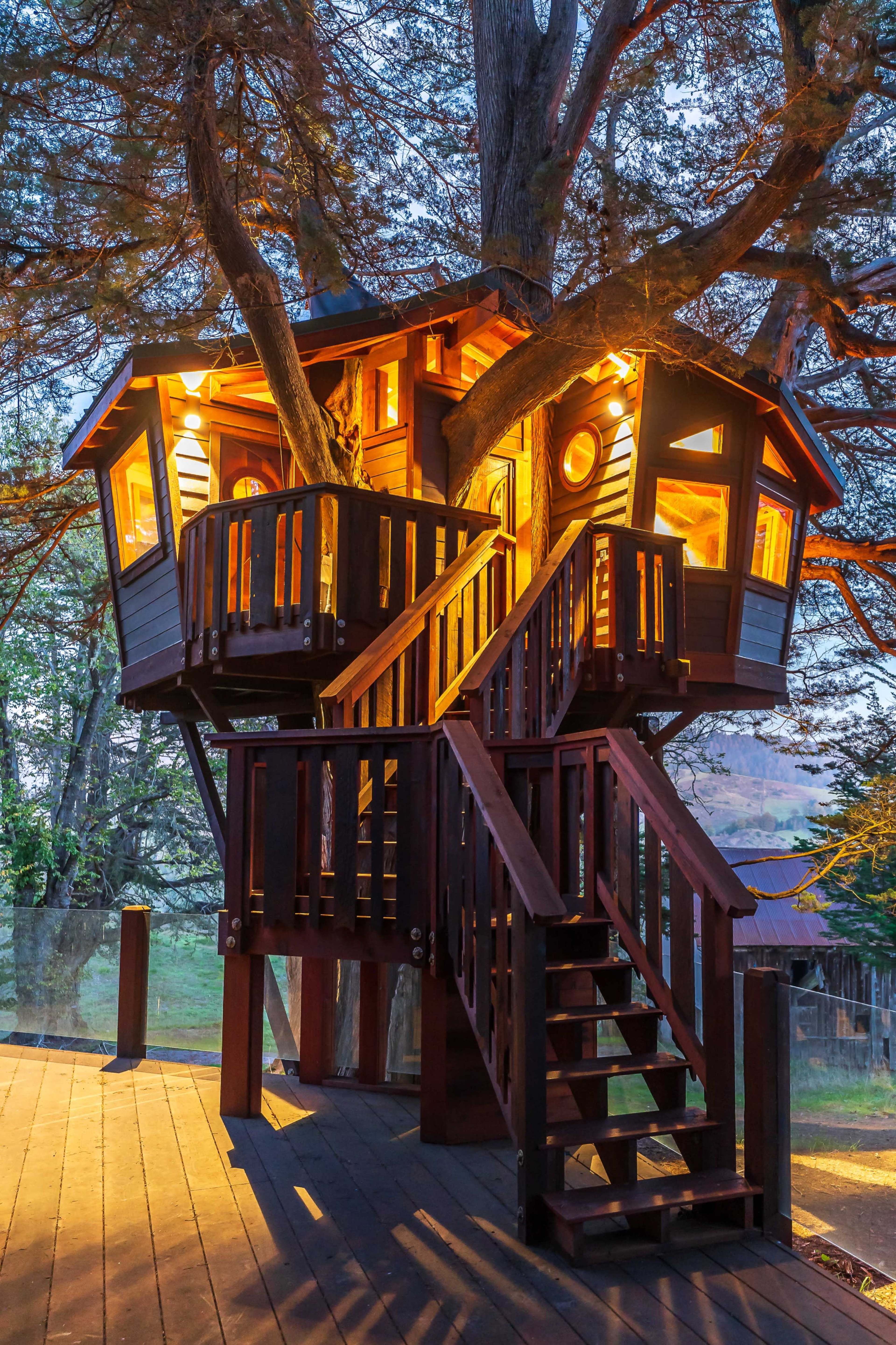 A wooden treehouse with multiple levels and warm lights illuminates its structure, nestled among the branches of a large tree.