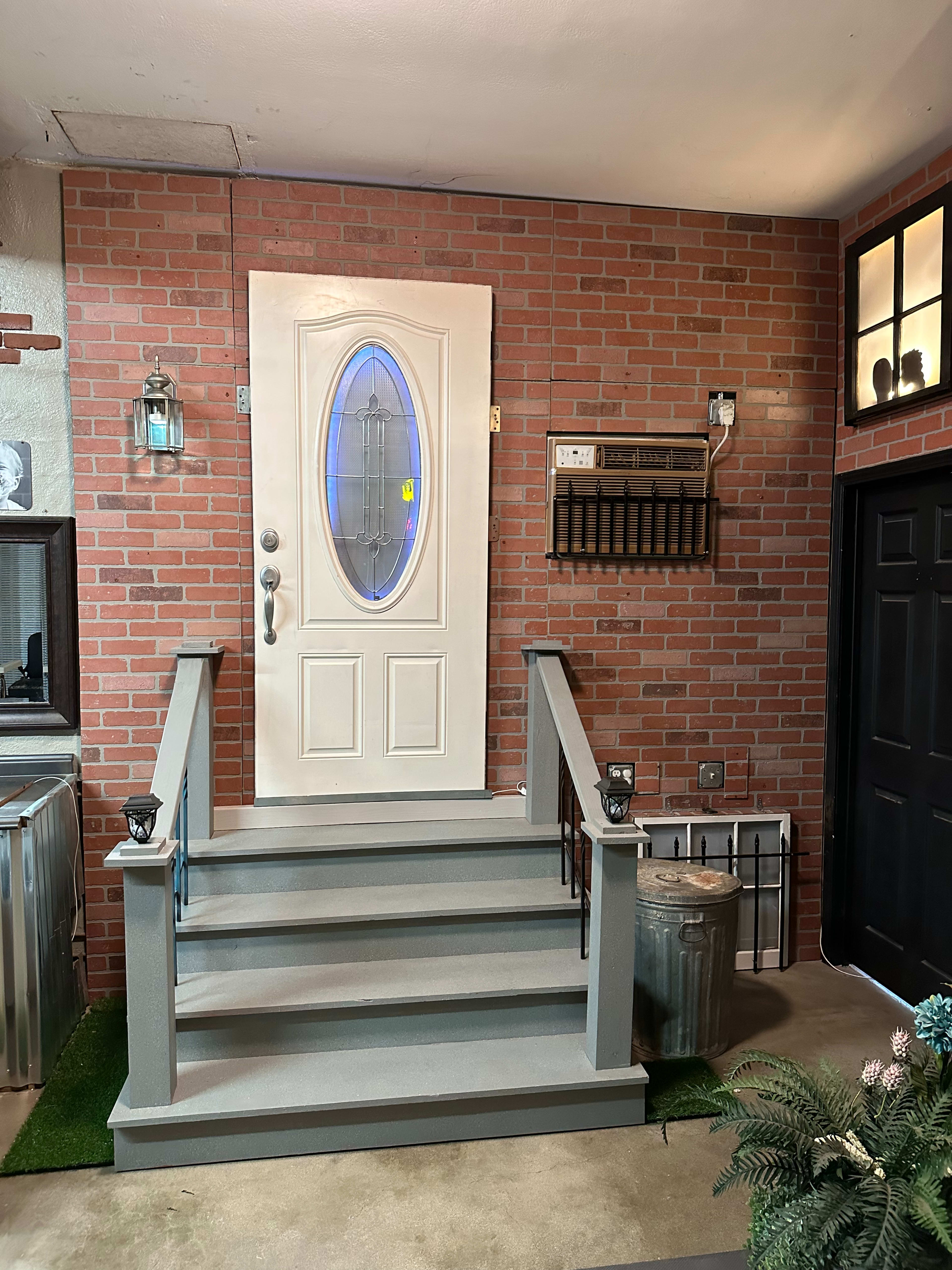 A white front door with an oval window, set against a brick wall, flanked by decorative lights and accompanied by steps leading to a small porch.
