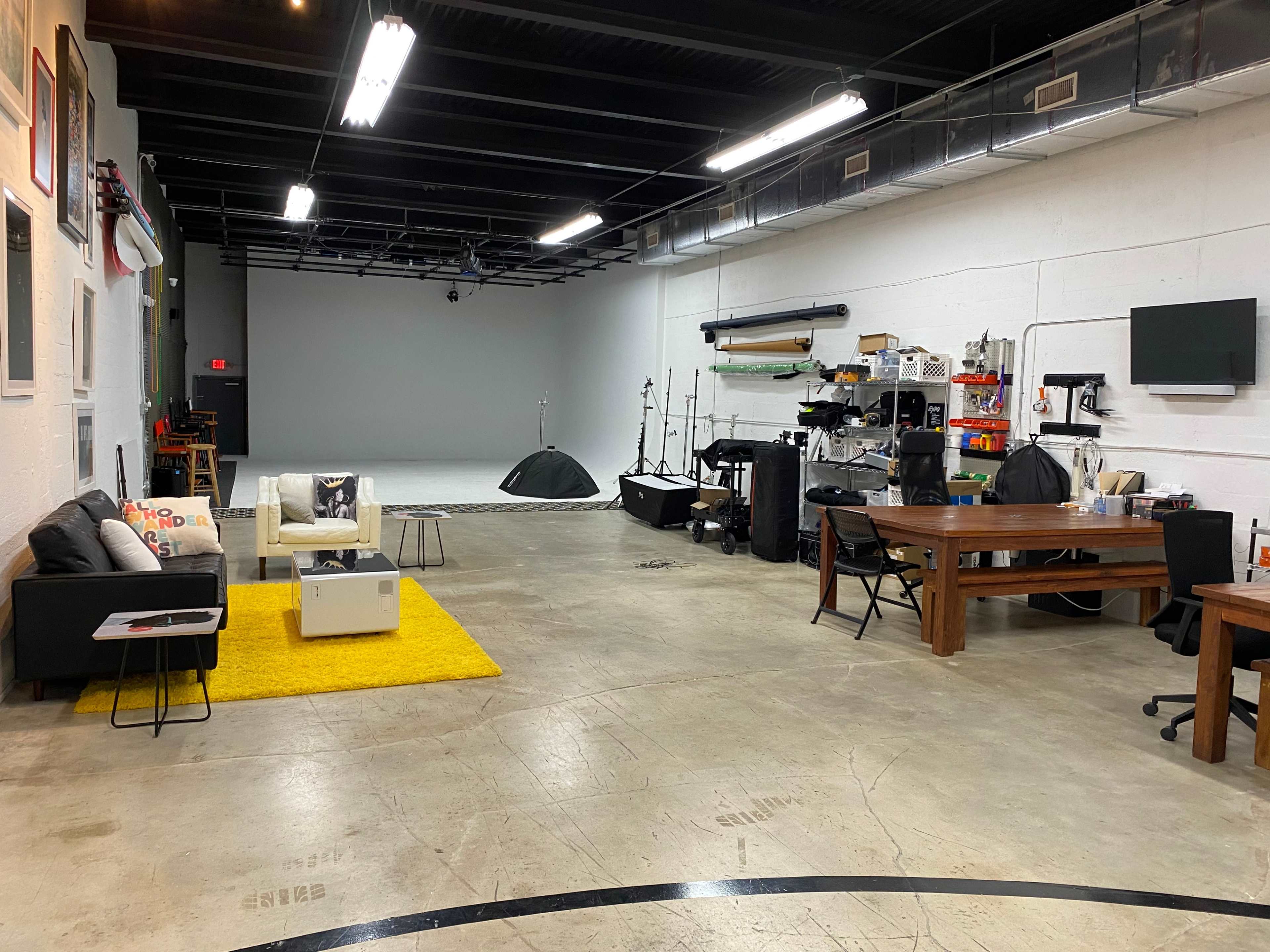 A spacious, well-lit studio featuring a minimalist seating area with a couch and yellow rug, alongside various equipment and furniture arranged throughout the concrete floor.