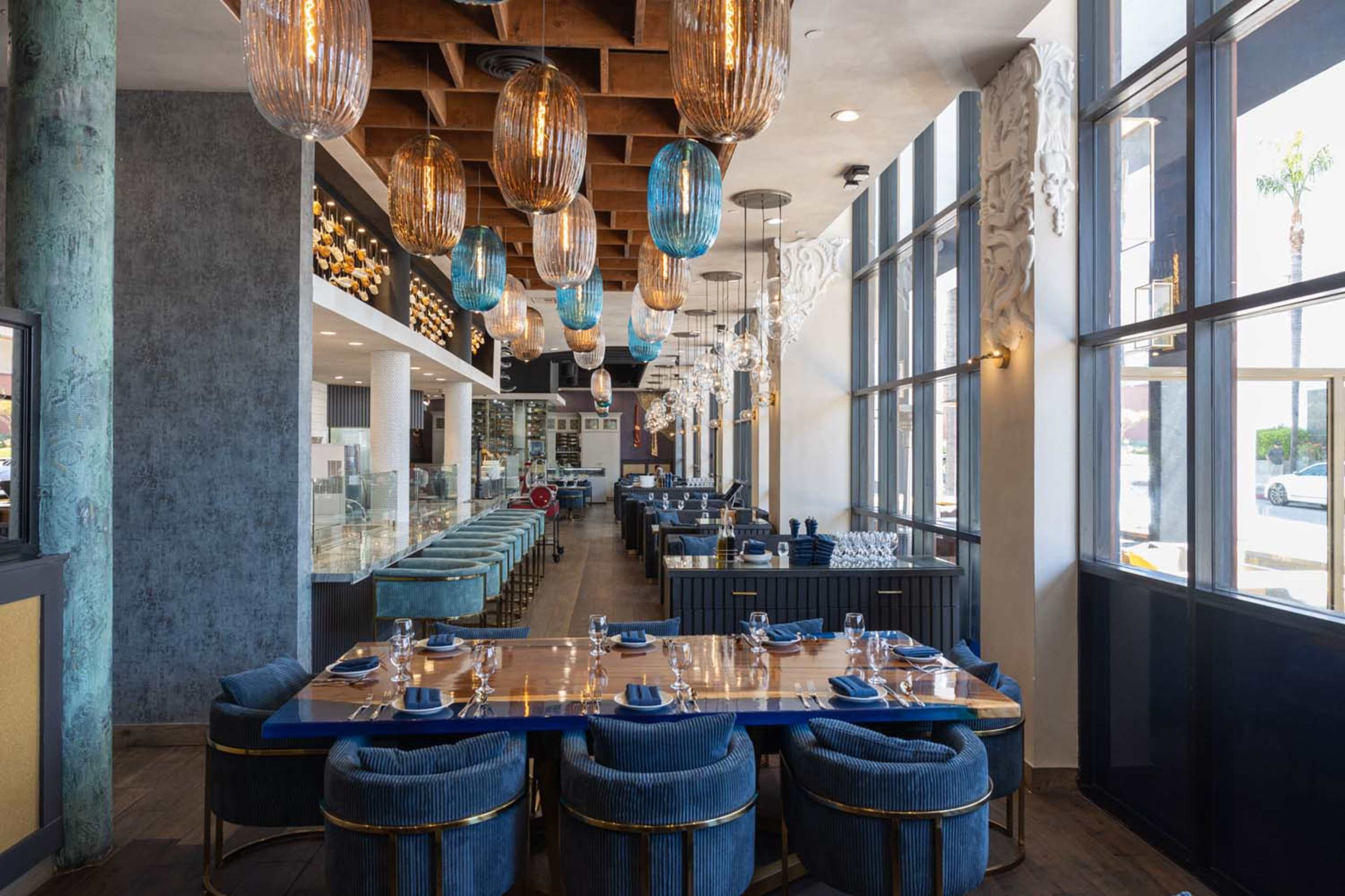 The image shows a modern restaurant interior with a long, blue upholstered dining table, wooden beams on the ceiling, and decorative pendant lighting.