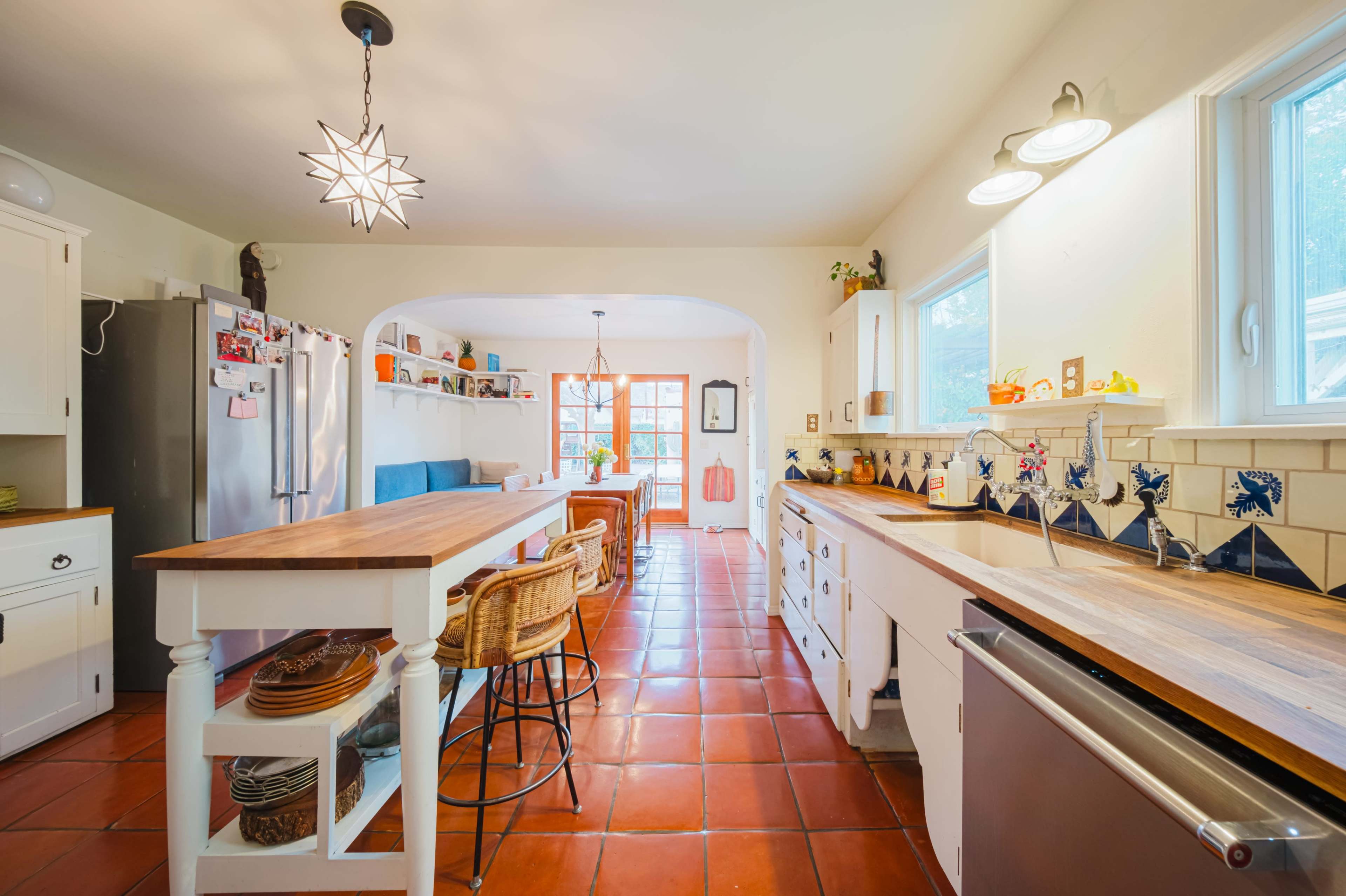 The image shows a bright kitchen featuring a wooden island, stainless steel appliances, and tile flooring, with a dining area visible in the background.