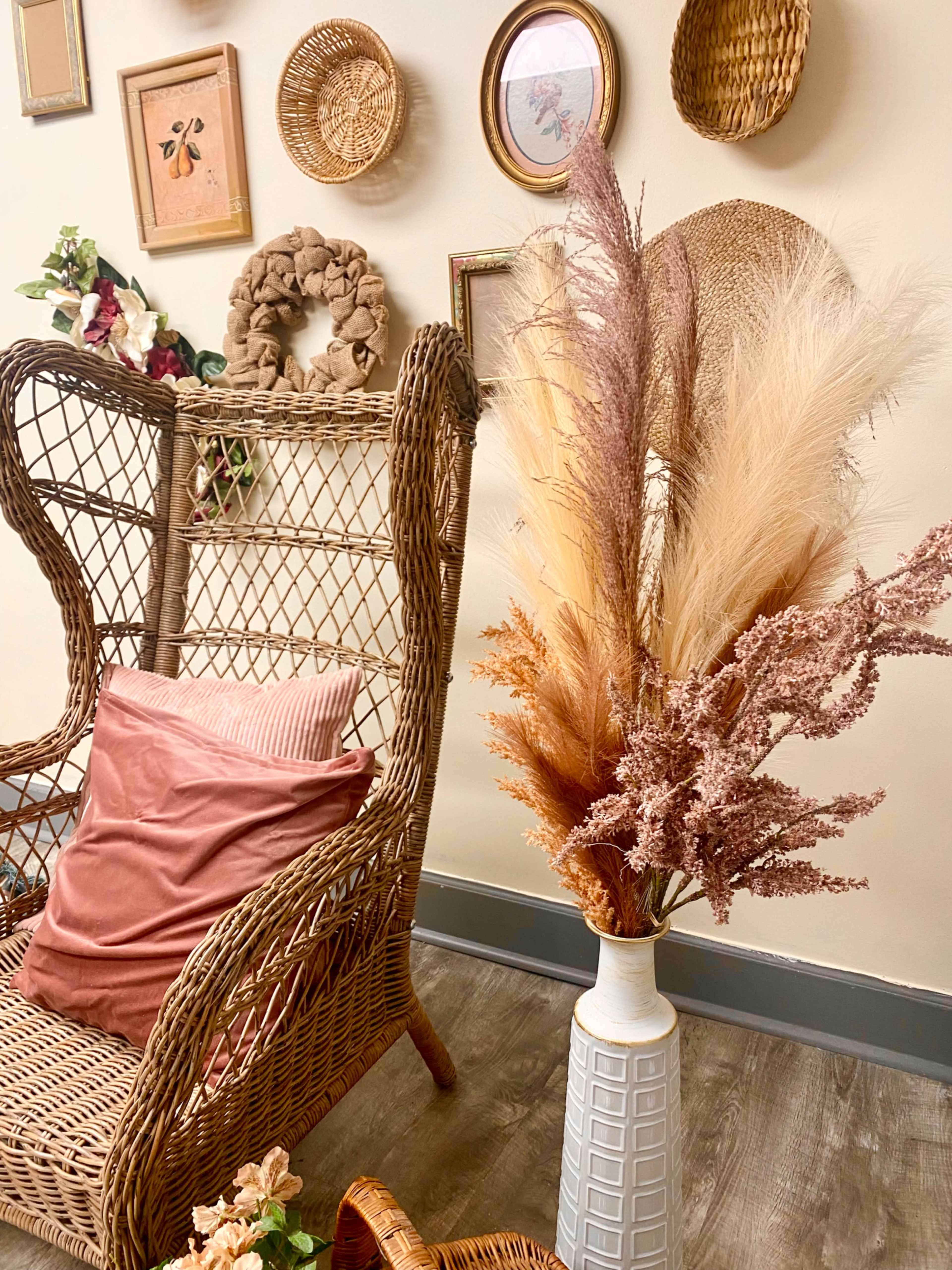 A wicker chair is positioned next to a vase filled with dried flowers, set against a wall adorned with various framed artworks and woven baskets.