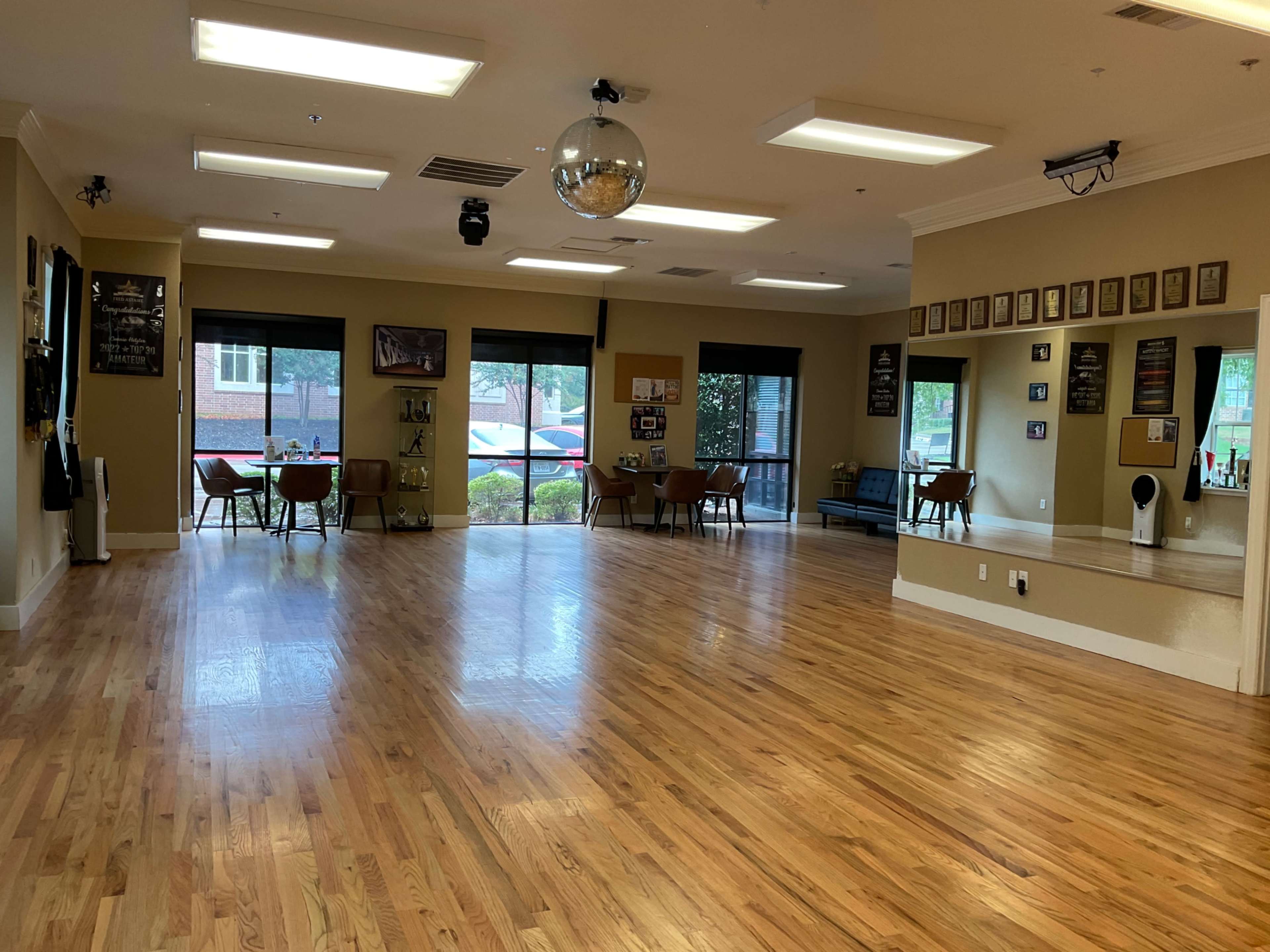 The image shows a spacious dance studio with wooden floors, large windows, and several tables and chairs arranged along one side.