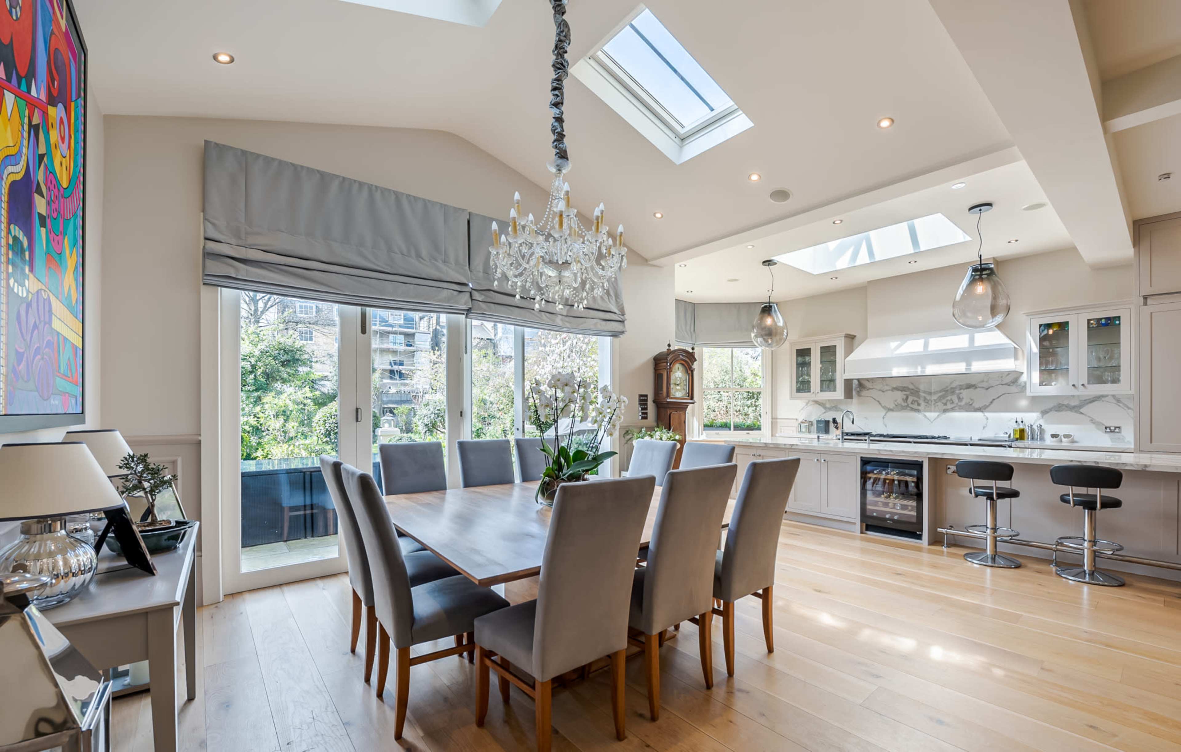 A modern kitchen and dining area featuring a large wooden dining table surrounded by grey upholstered chairs, with large windows and skylights providing natural light.