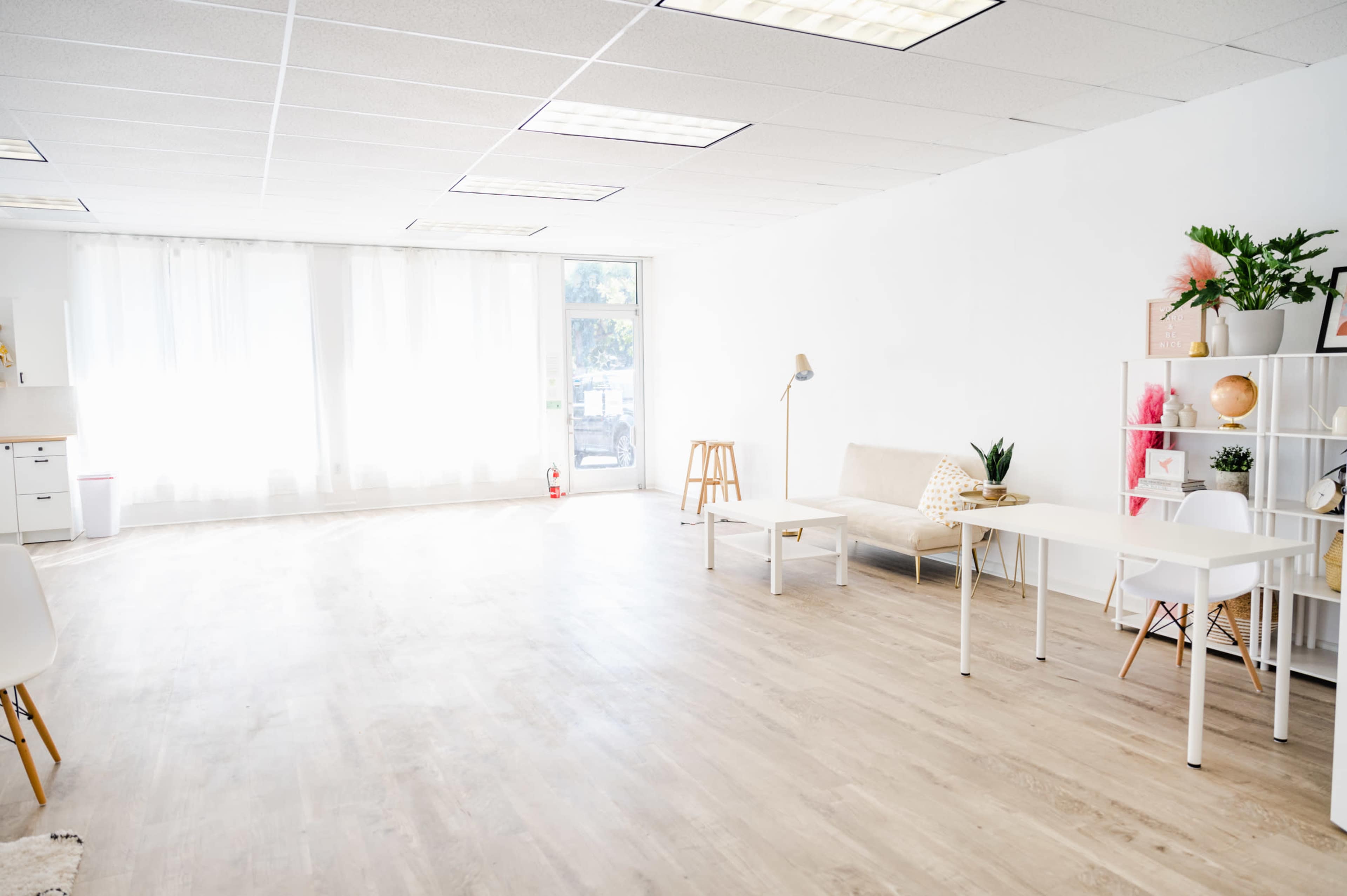 The image shows a bright, minimalist room with light-colored wooden flooring, large windows, a sofa, a desk, and decorative plants.