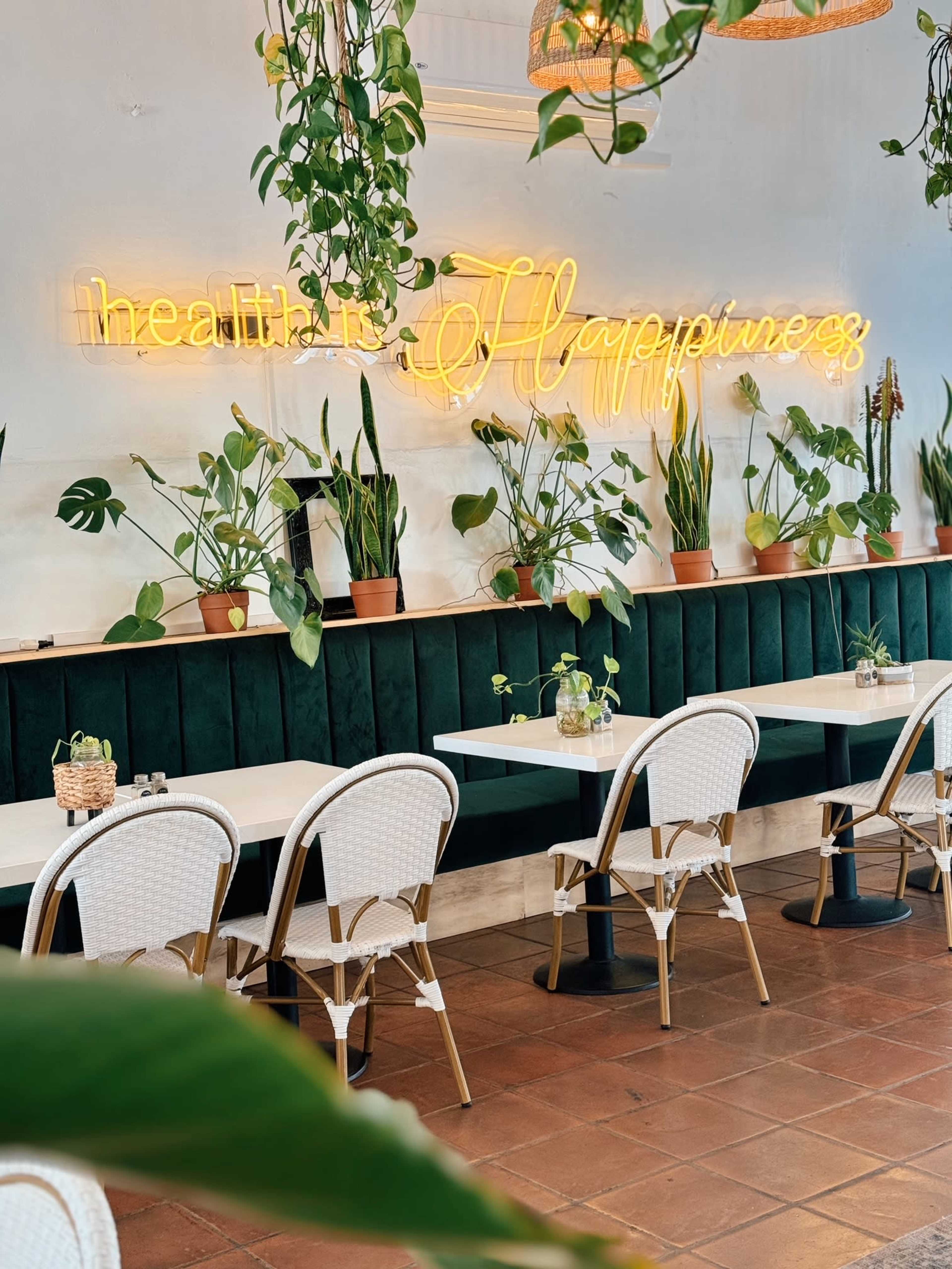 A stylish cafe interior with green velvet seating, light wooden tables, and potted plants, featuring a neon sign that reads "health is Happiness."