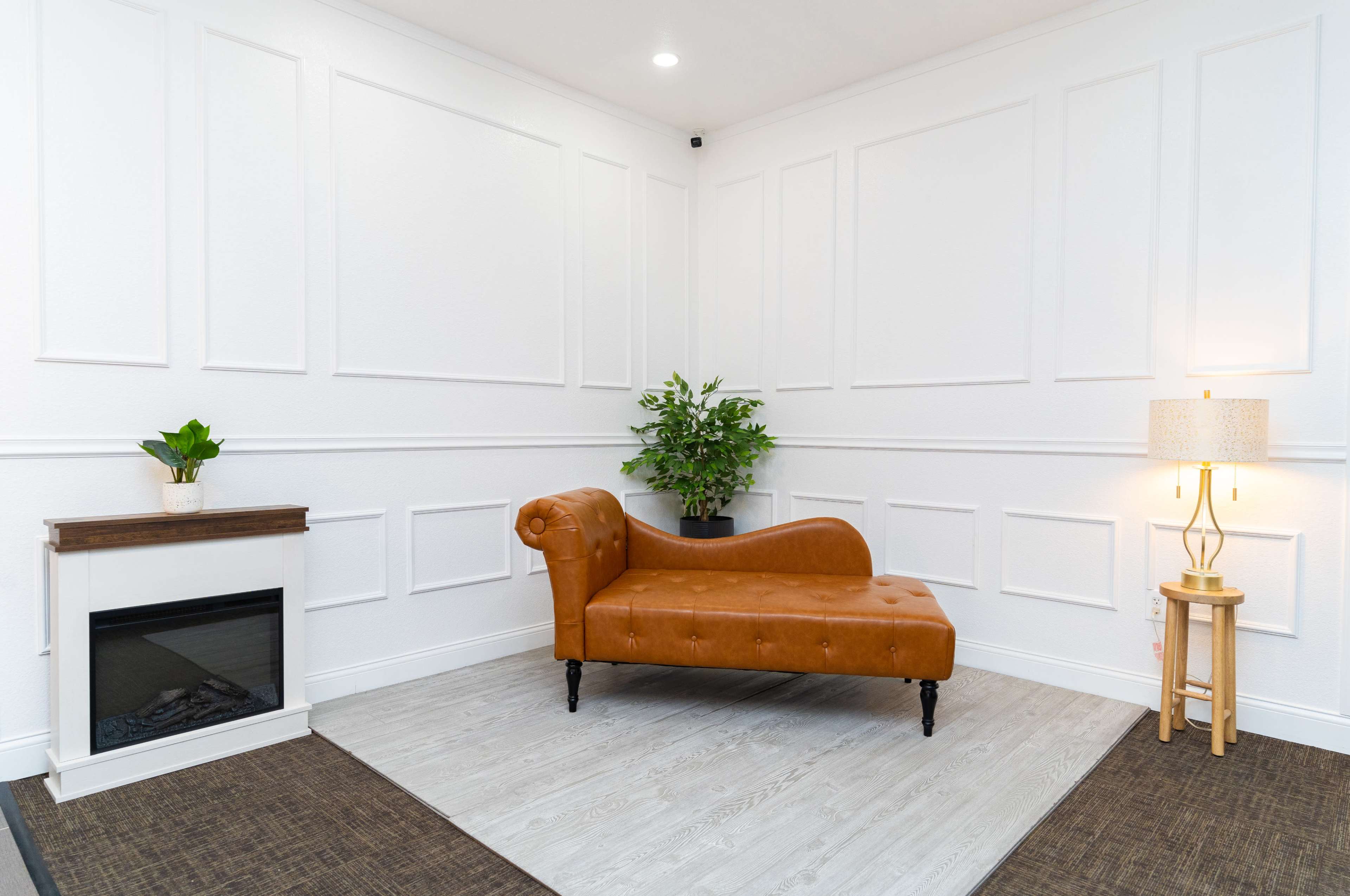 The image features a cozy interior with a brown chaise lounge, a potted plant, and a floor lamp beside a small fireplace against a white paneled wall.