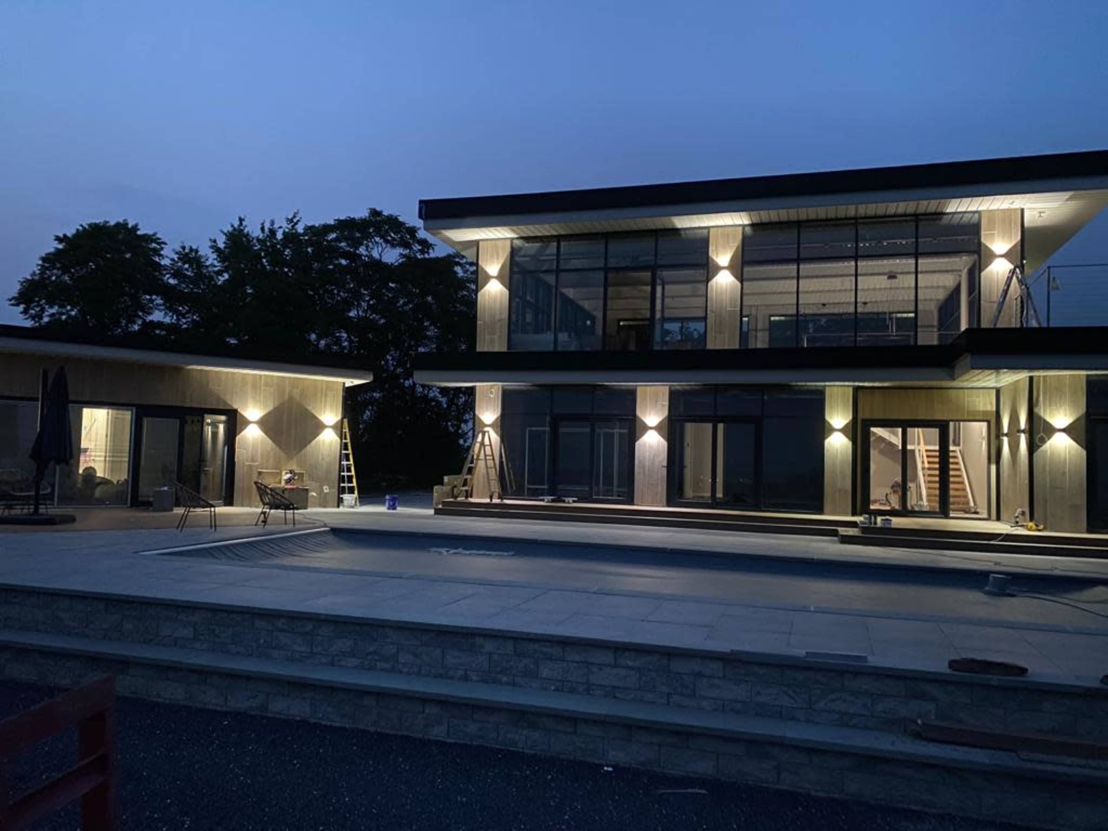 A modern two-story house with large windows is illuminated by artificial lights at dusk, featuring a surrounding patio and pool area.