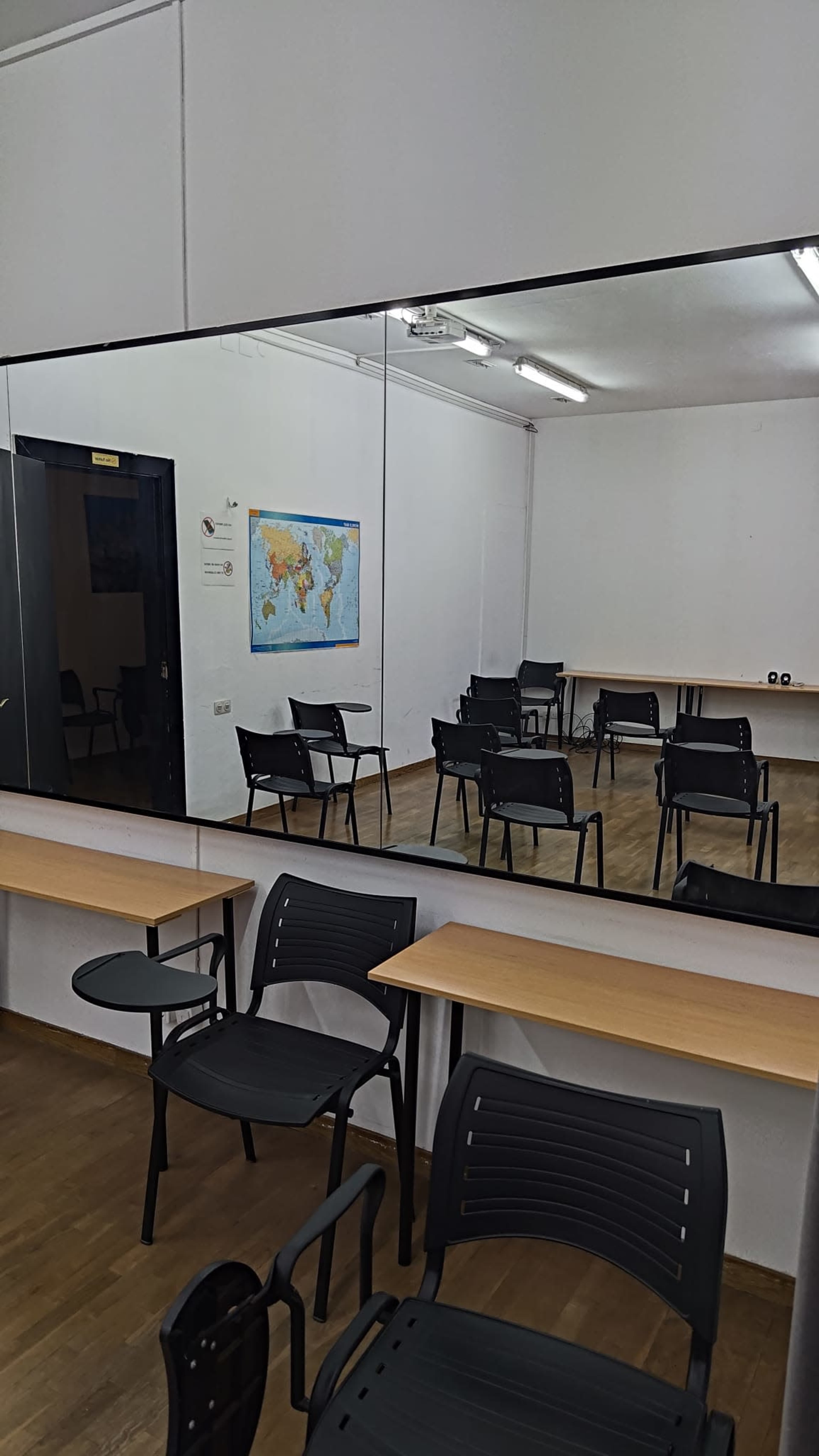 The image shows a classroom with several black chairs arranged around a wooden table and a world map hanging on the wall, reflected in a large mirror.