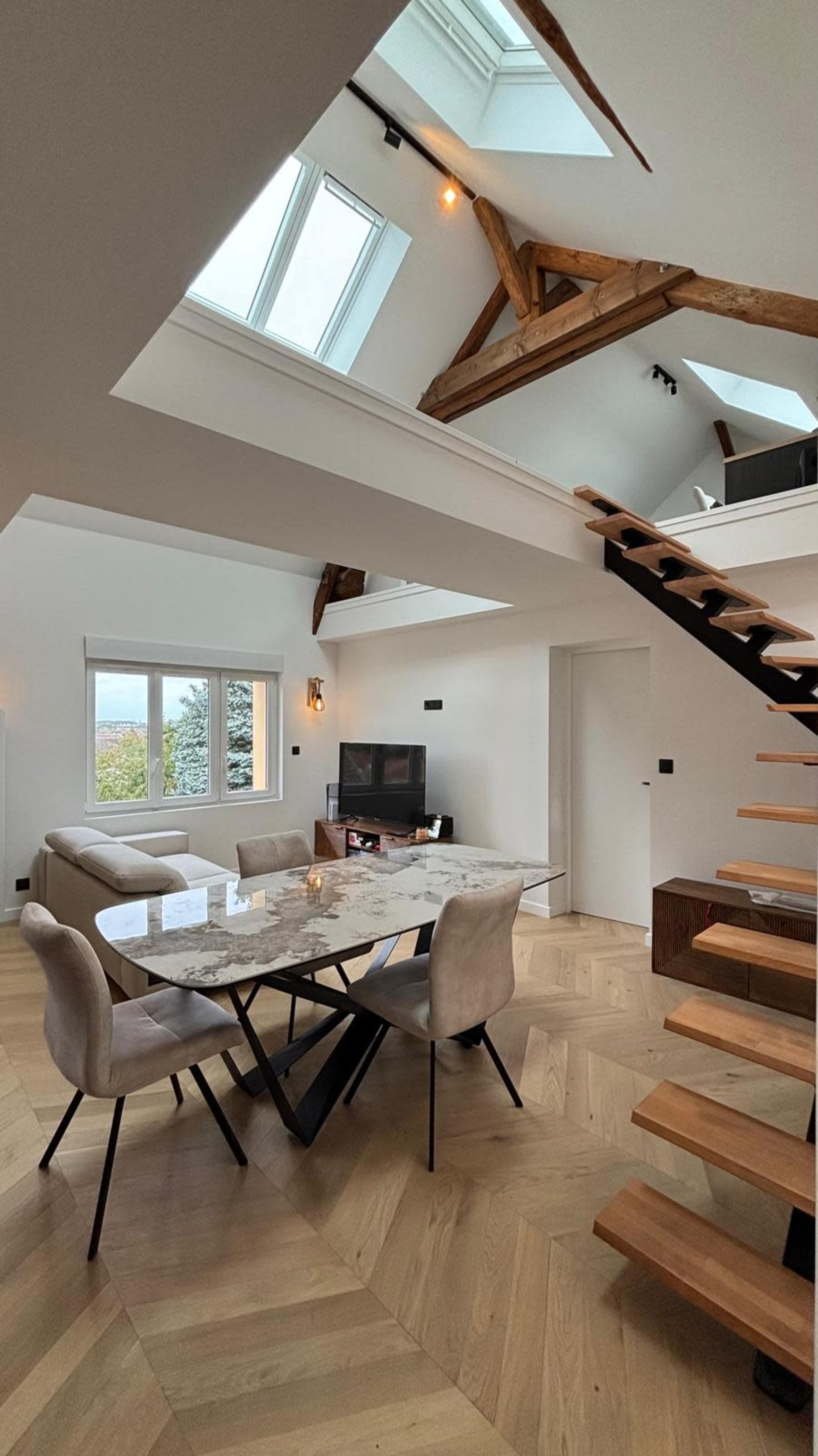 The image shows a modern living area featuring a dining table with four chairs, a sofa, a television, and a staircase leading to an upper level beneath a skylight.