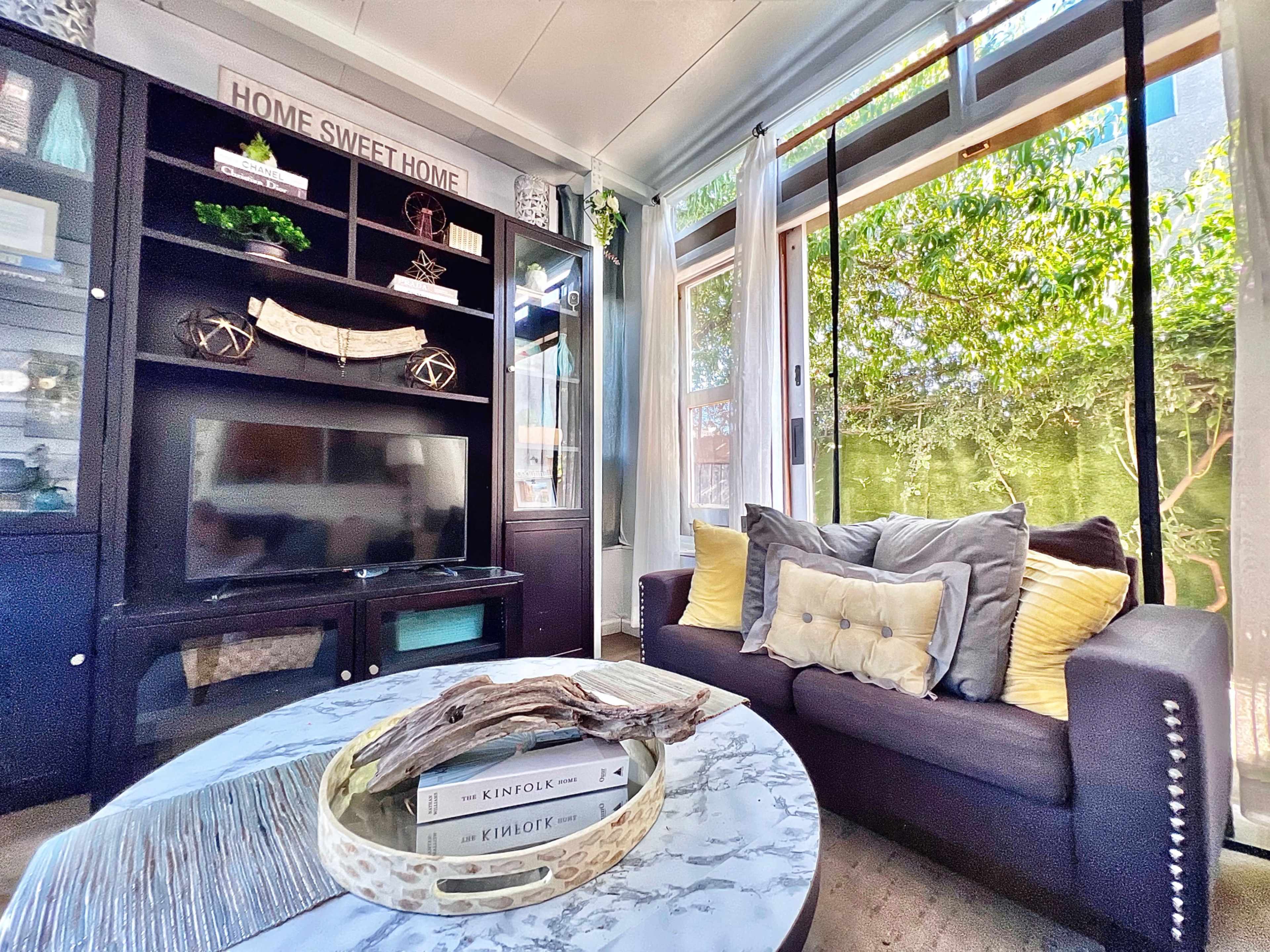 A cozy living room features a dark cabinet with a television, a round marble coffee table with books, and a sofa adorned with decorative pillows, all set against a backdrop of large windows letting in natural light and greenery.