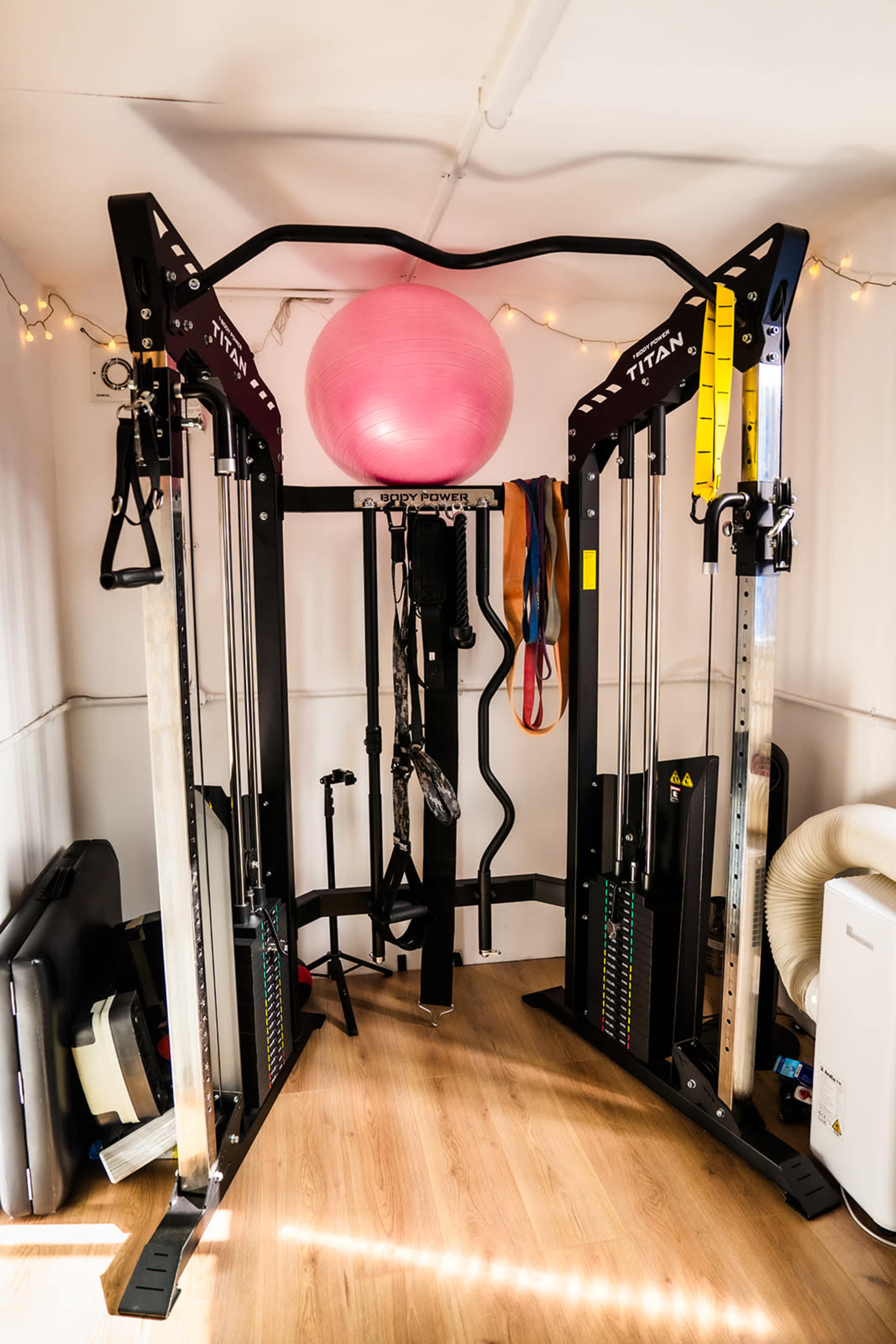 The image shows a home gym setup featuring a cable machine with various attachments, a large exercise ball, and resistance bands in a brightly lit room with wooden flooring.