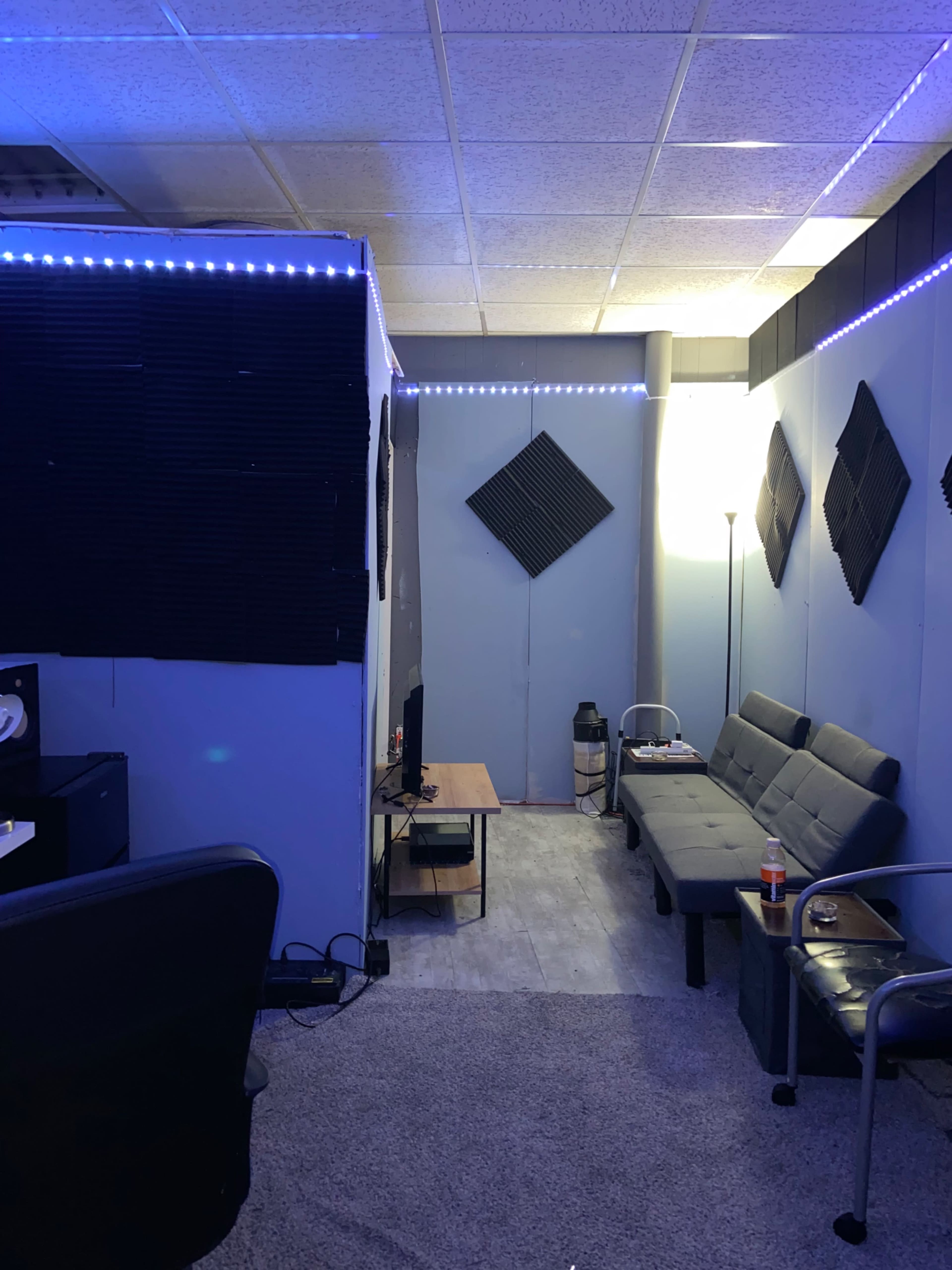 A small recording studio with blue walls, acoustic panels, a couch, a coffee table, and ambient lighting along the ceiling.