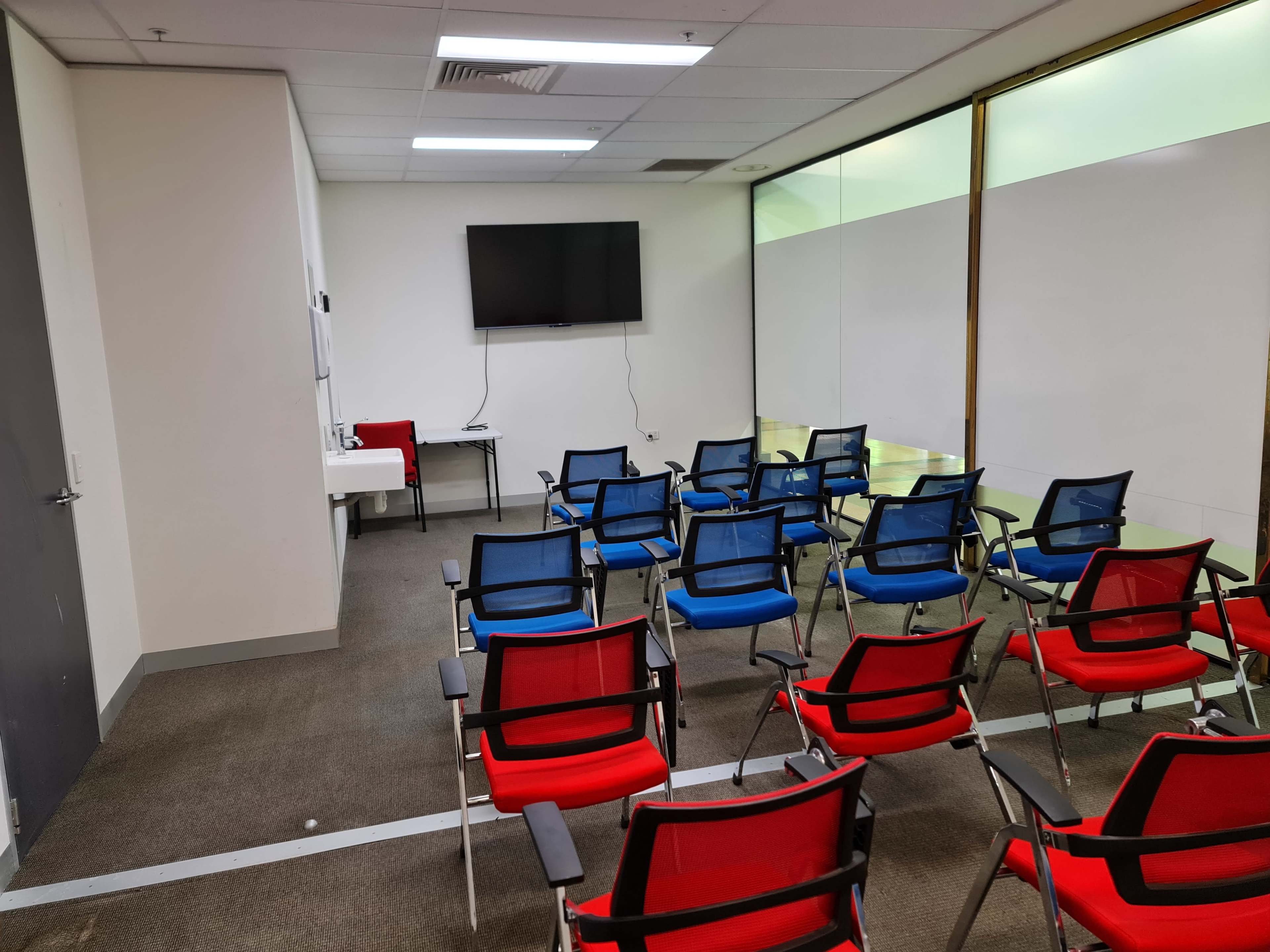 The image shows a meeting room with red and blue chairs arranged in rows facing a wall-mounted television and a table.