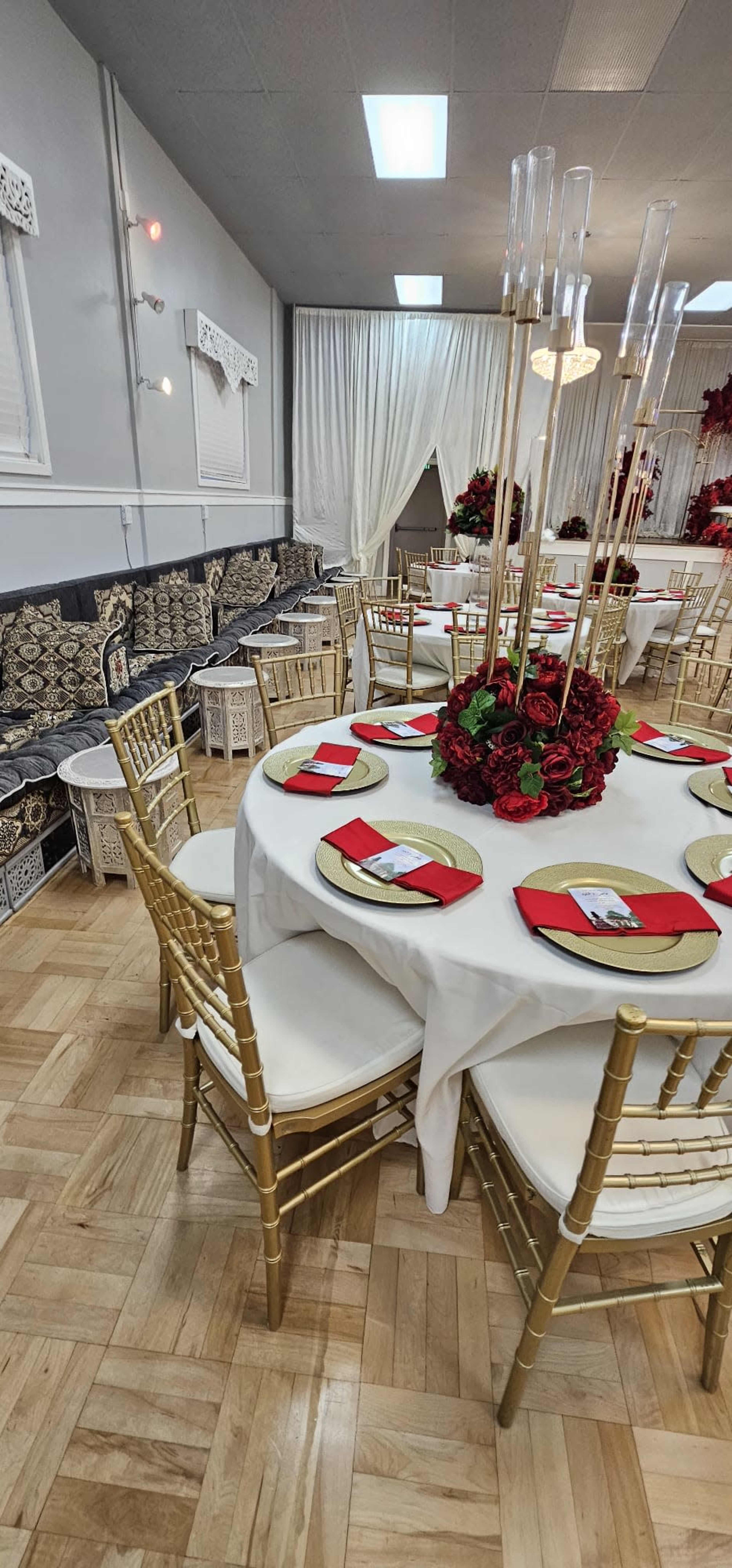 A banquet hall is set up with round tables covered in white linen, adorned with floral centerpieces and gold chiavari chairs.