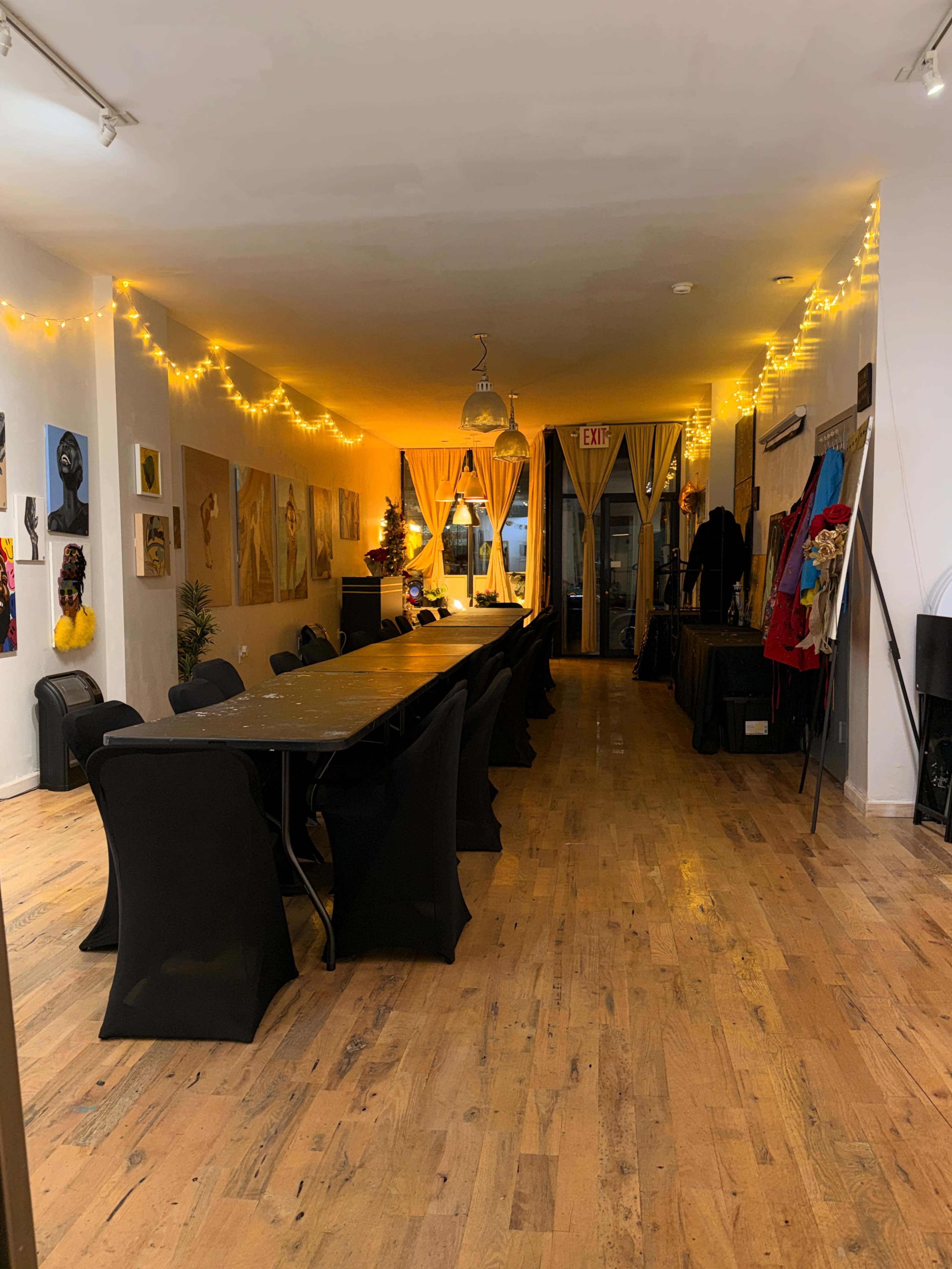 The image shows a long, narrow room with a large table surrounded by black chairs, illuminated by string lights and featuring artwork on the walls.