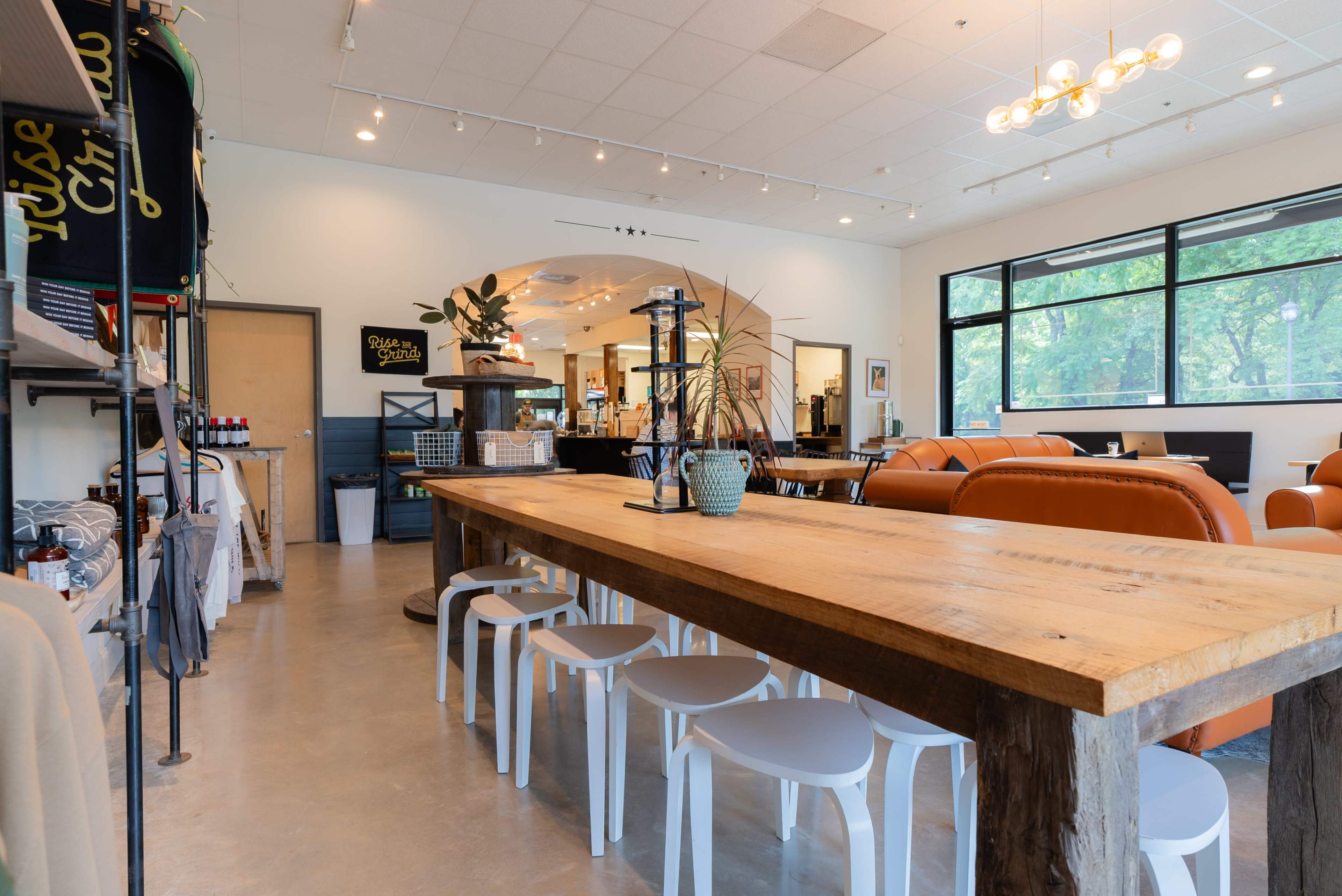 The image shows a modern café interior with a large wooden communal table surrounded by white chairs, orange seating areas, and large windows letting in natural light.