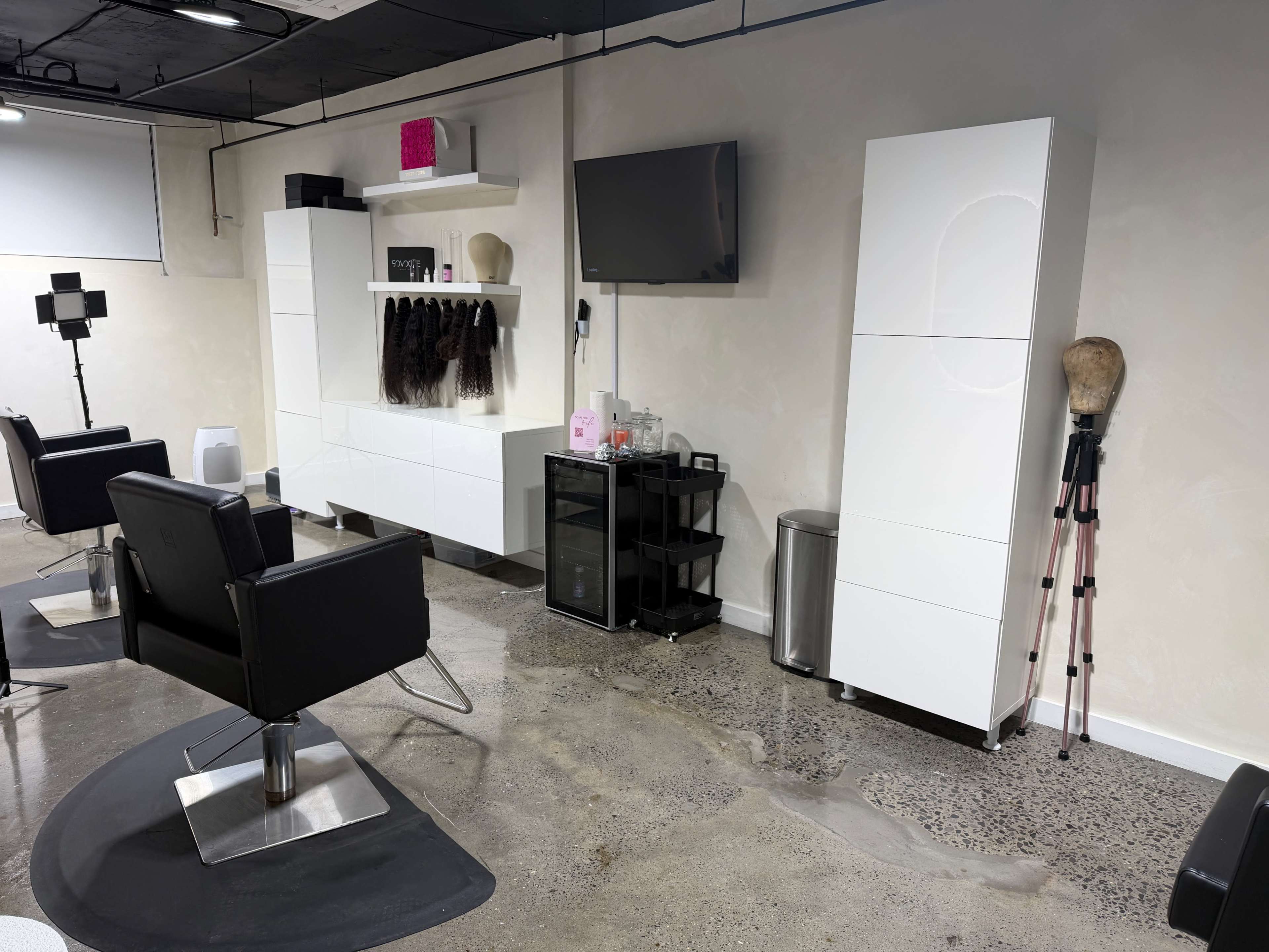 The image shows a modern hairstyling salon featuring black chairs, white cabinetry, a television, and a mannequin head on a stand.