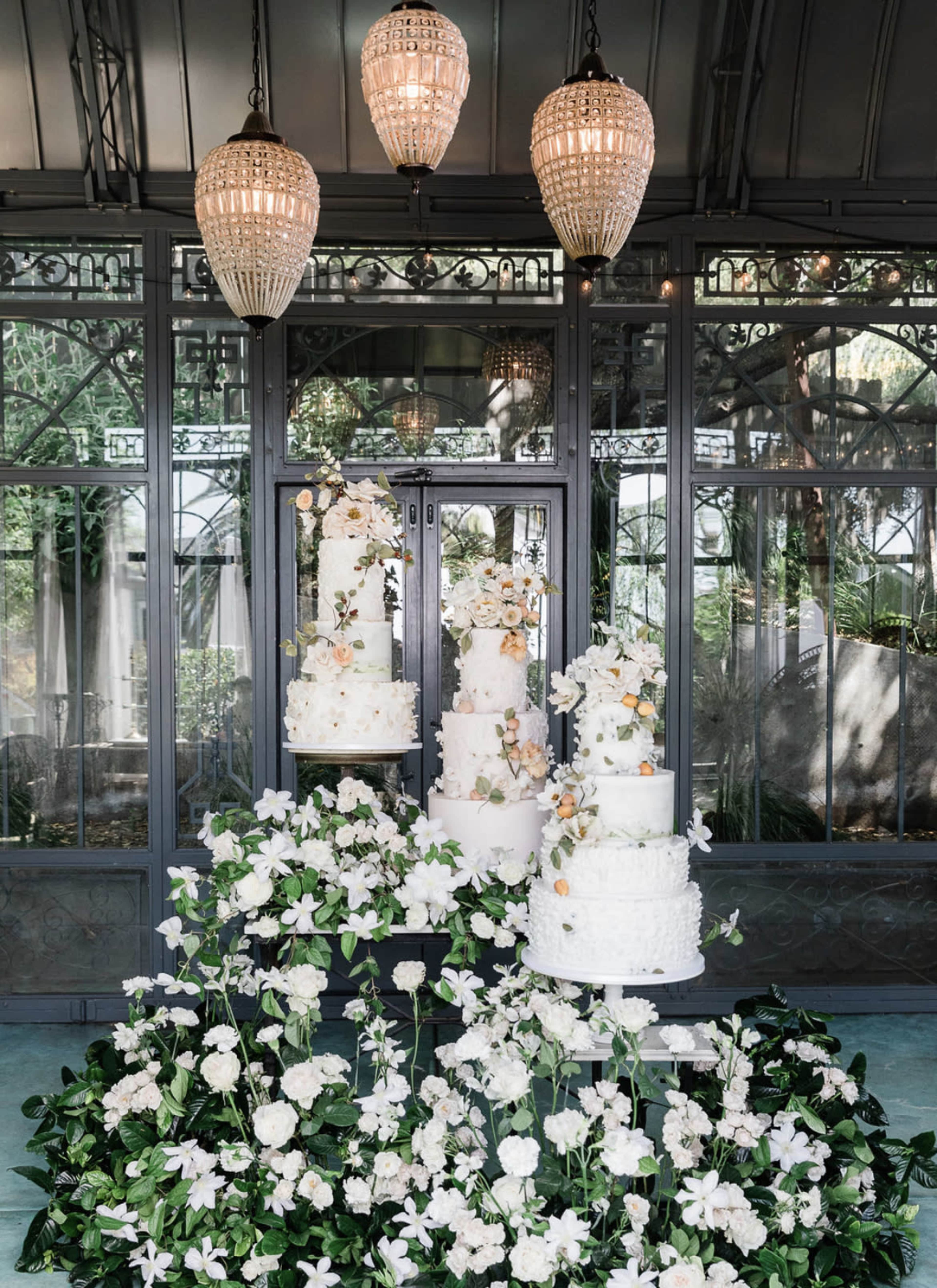 A display of several elegant white wedding cakes adorned with flowers stands on a pedestal surrounded by greenery in a beautifully designed indoor space.
