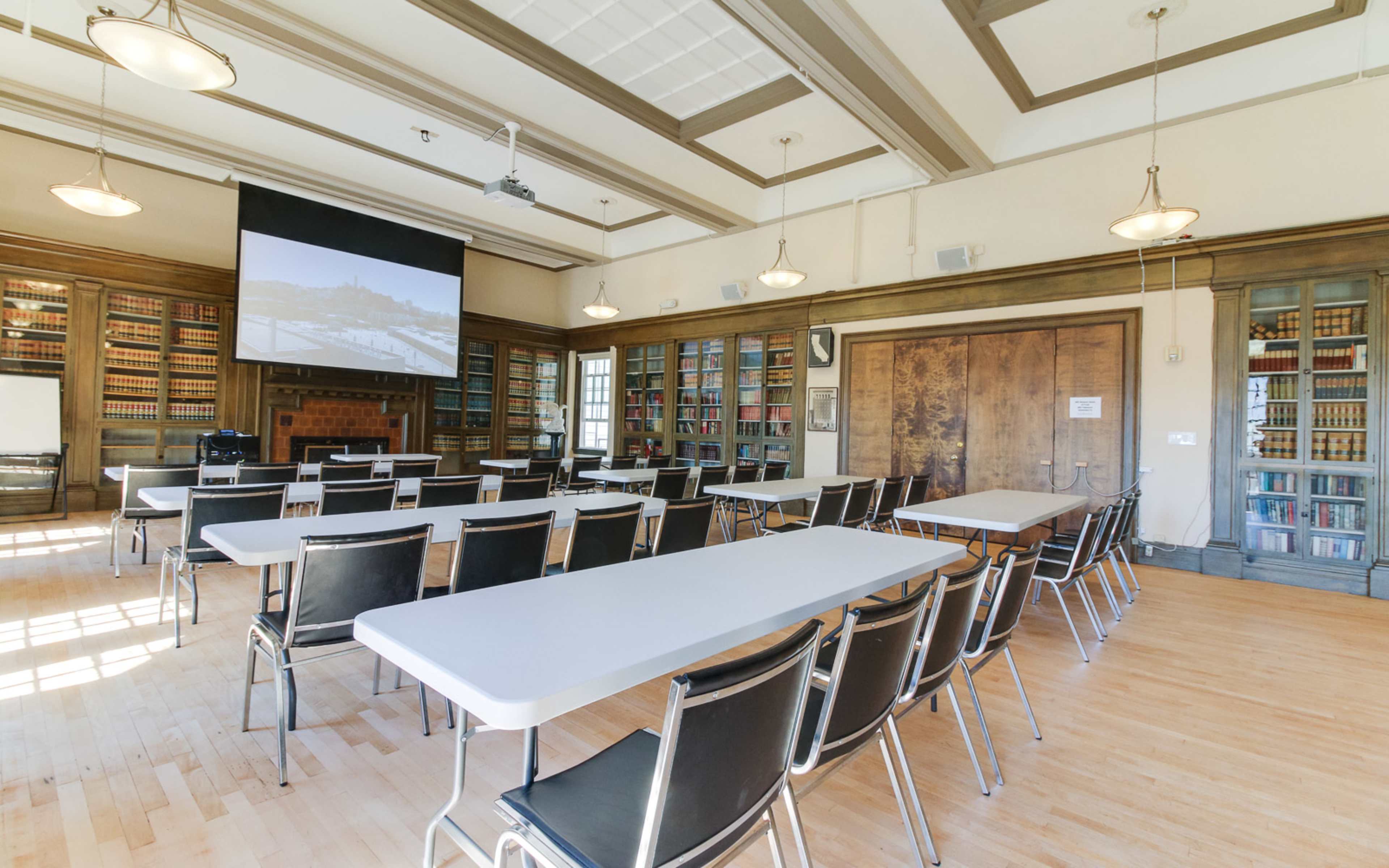 The image shows a spacious classroom with multiple tables and chairs arranged in rows, along with a projector screen at one end and bookshelves lining the walls.