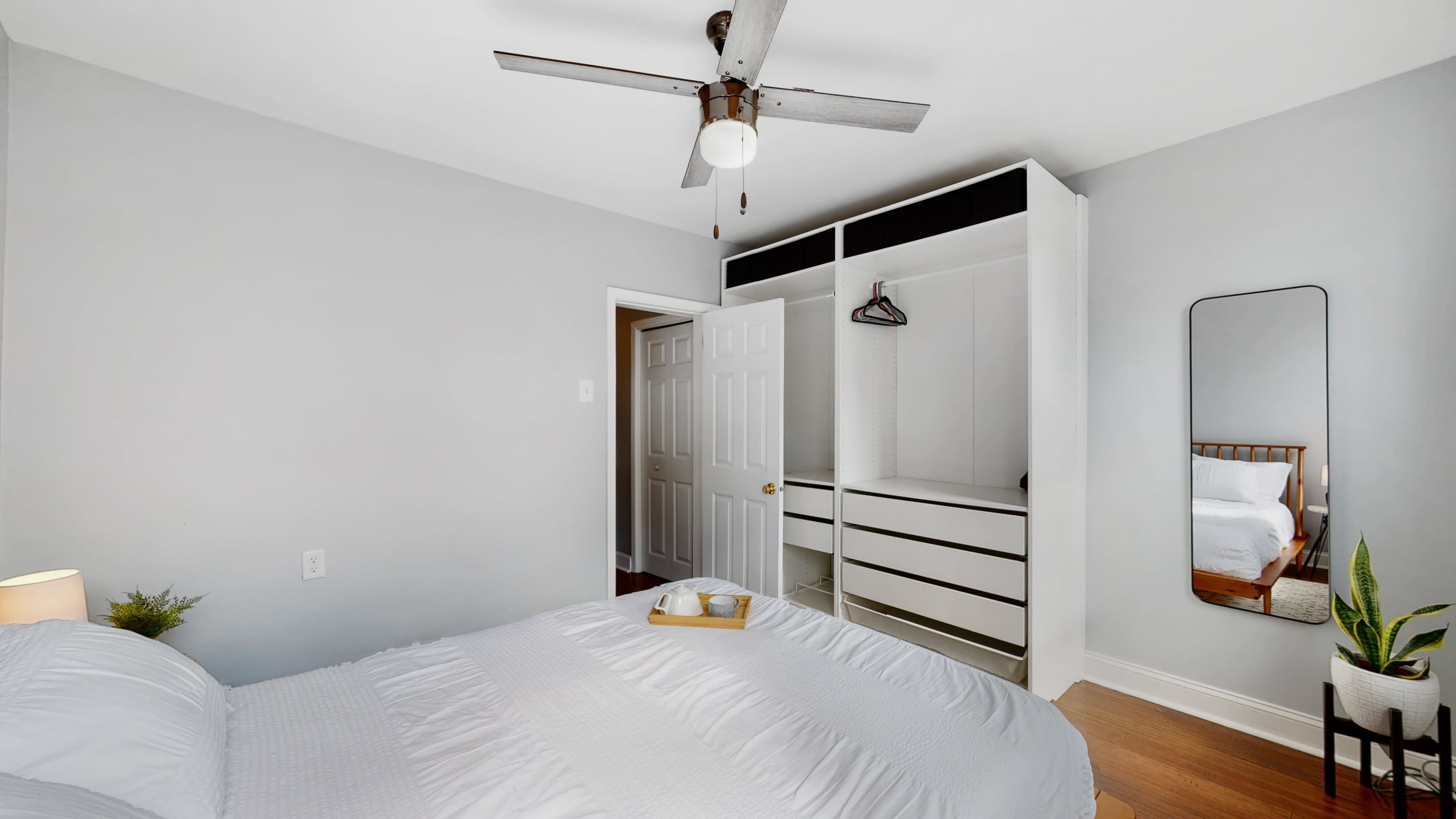 A neatly arranged bedroom features a bed with a tray on top, a ceiling fan, a closet with hanging space, and a mirror reflecting the room's interior.