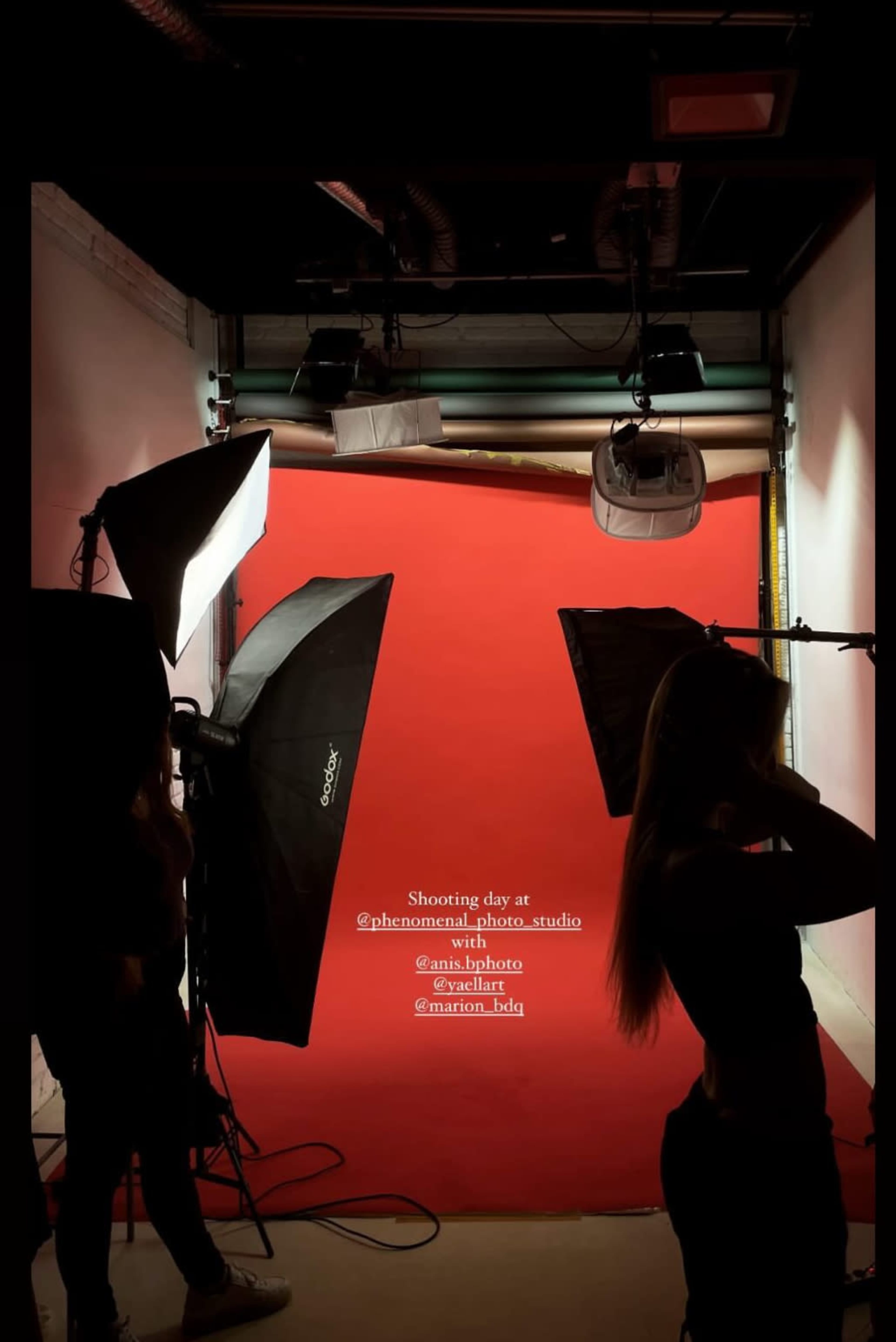 A silhouette of a model poses against a red backdrop in a photography studio, surrounded by lighting equipment.
