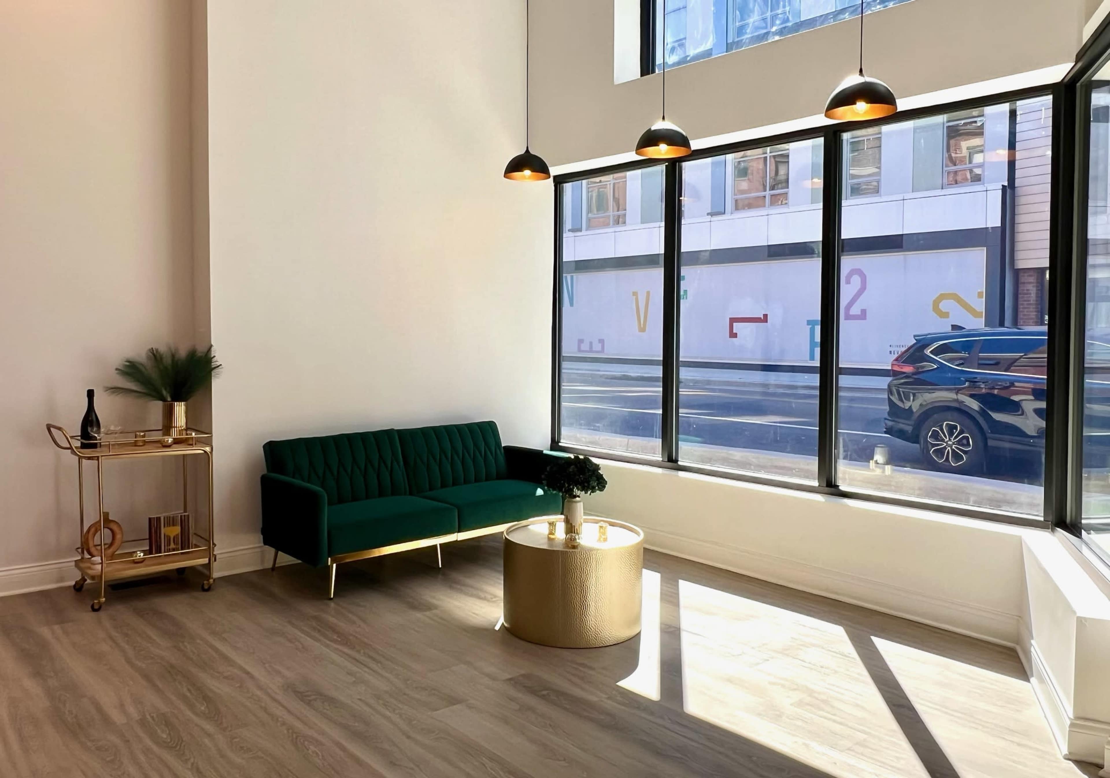 A green sofa and a gold coffee table sit in a well-lit room with large windows overlooking a street.