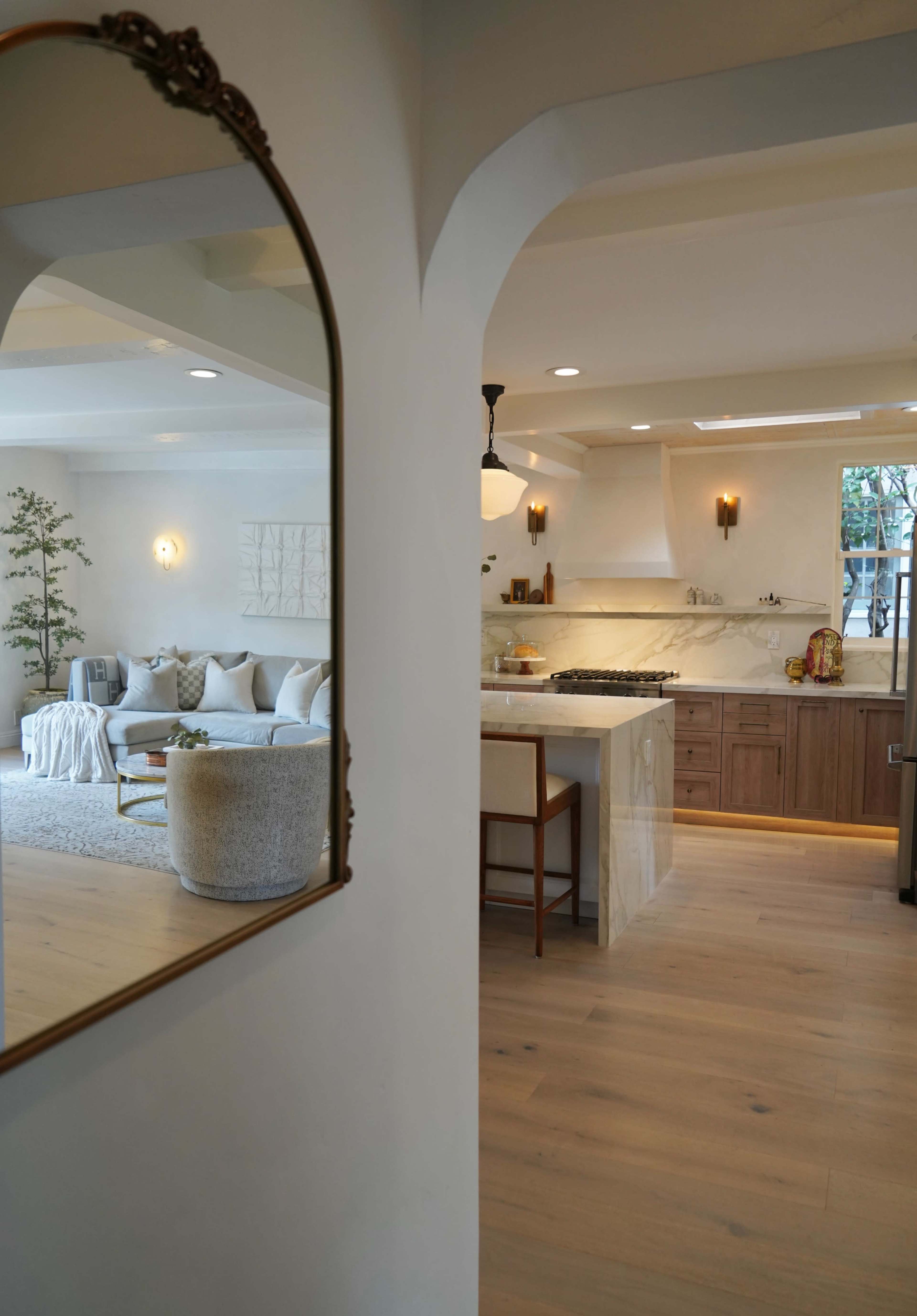 The image shows a modern, open-concept living space with a kitchen visible through an arched mirror frame.