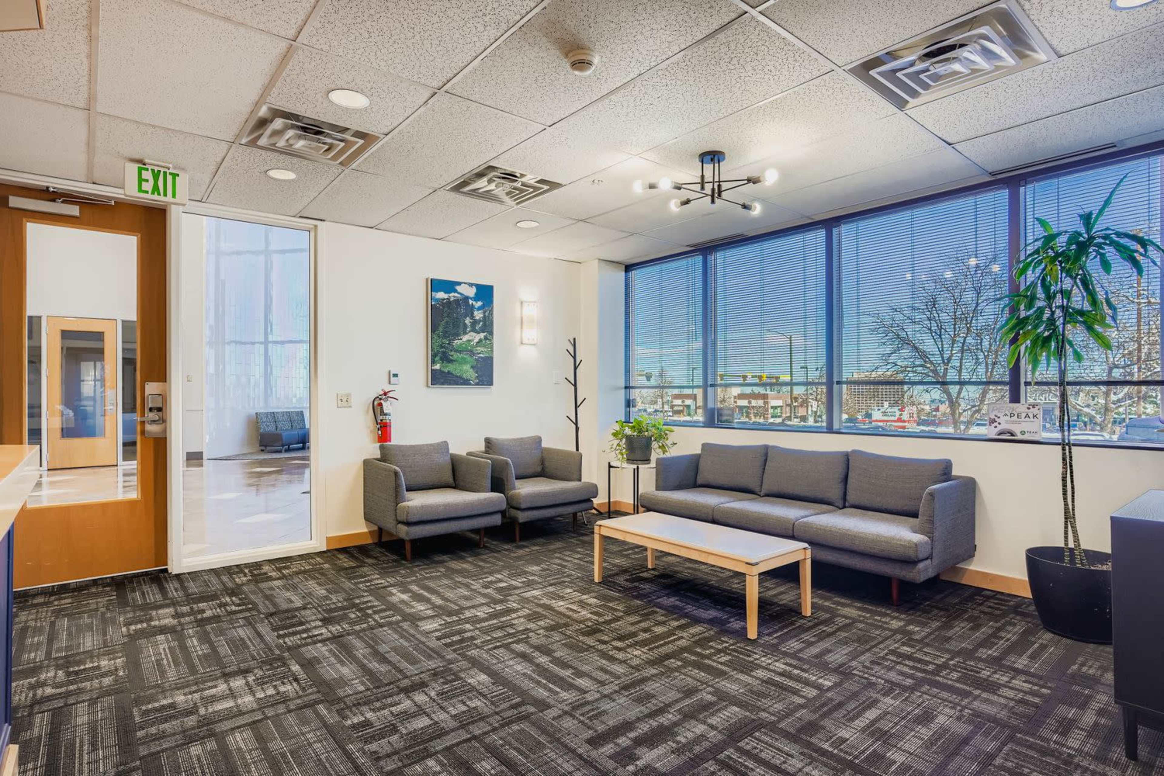 The image shows a modern office lounge area with gray sofas, a coffee table, large windows, and a potted plant.