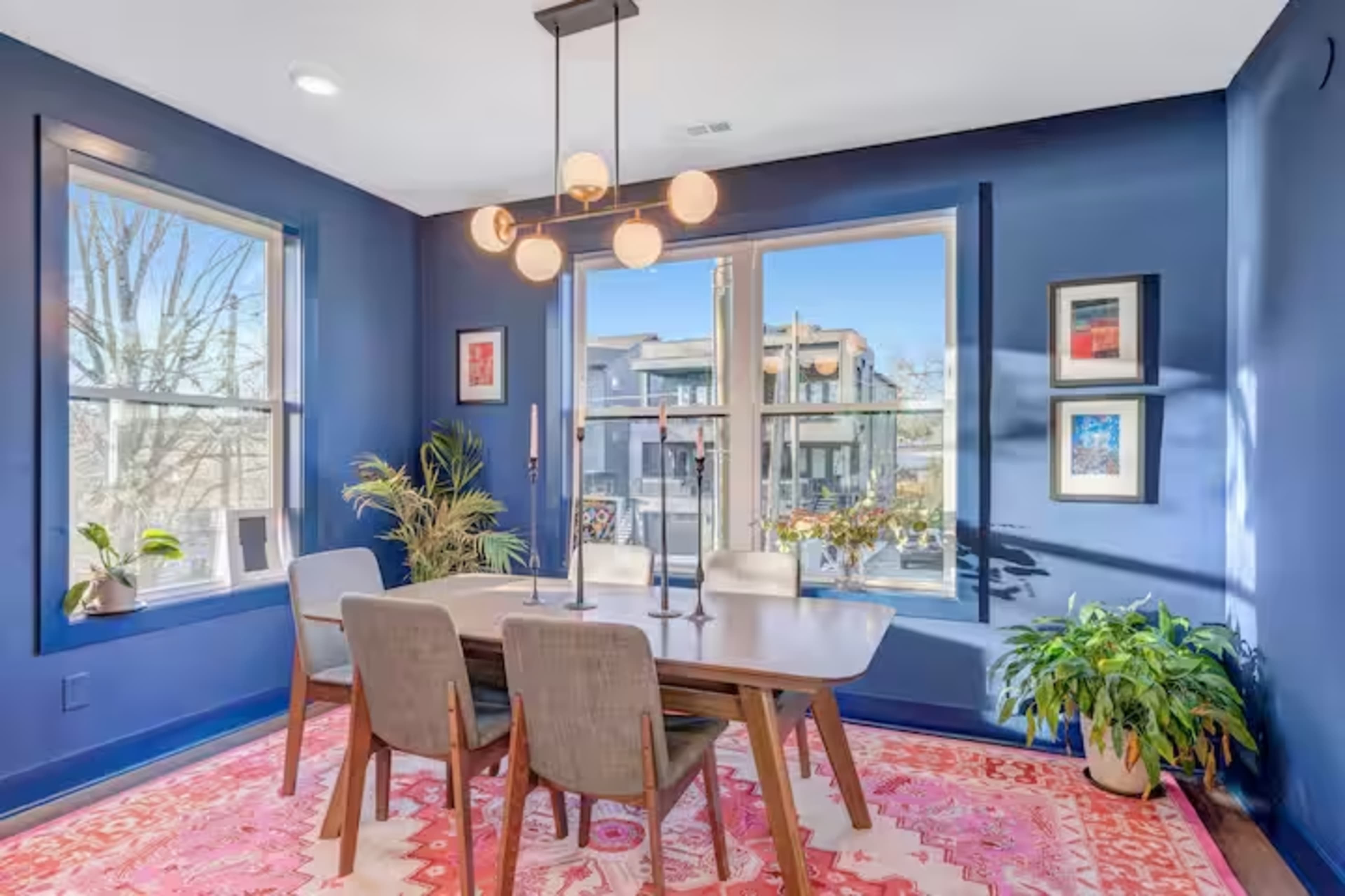 A dining area with a wooden table and four chairs is brightly lit by natural light from large windows, set against deep blue walls and decorated with framed art and indoor plants.