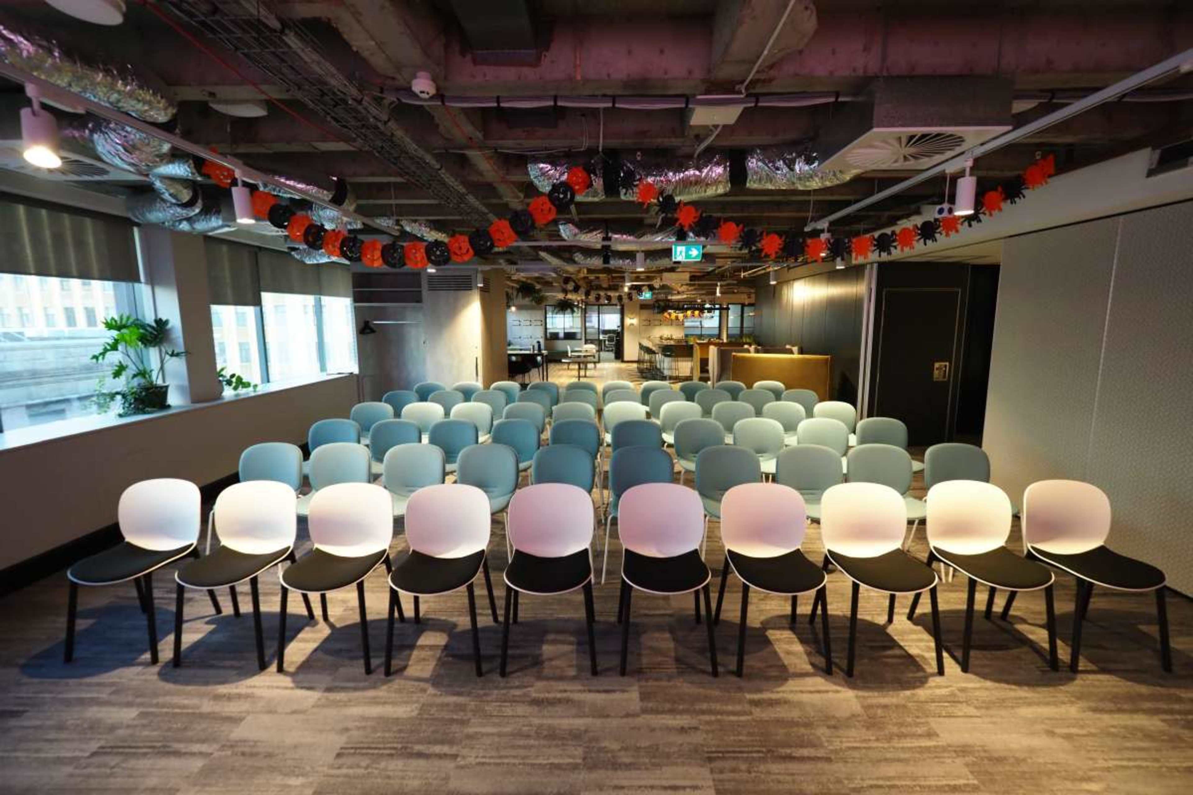 The image shows a bright, modern meeting room arranged with rows of light blue and black chairs facing a presentation area.