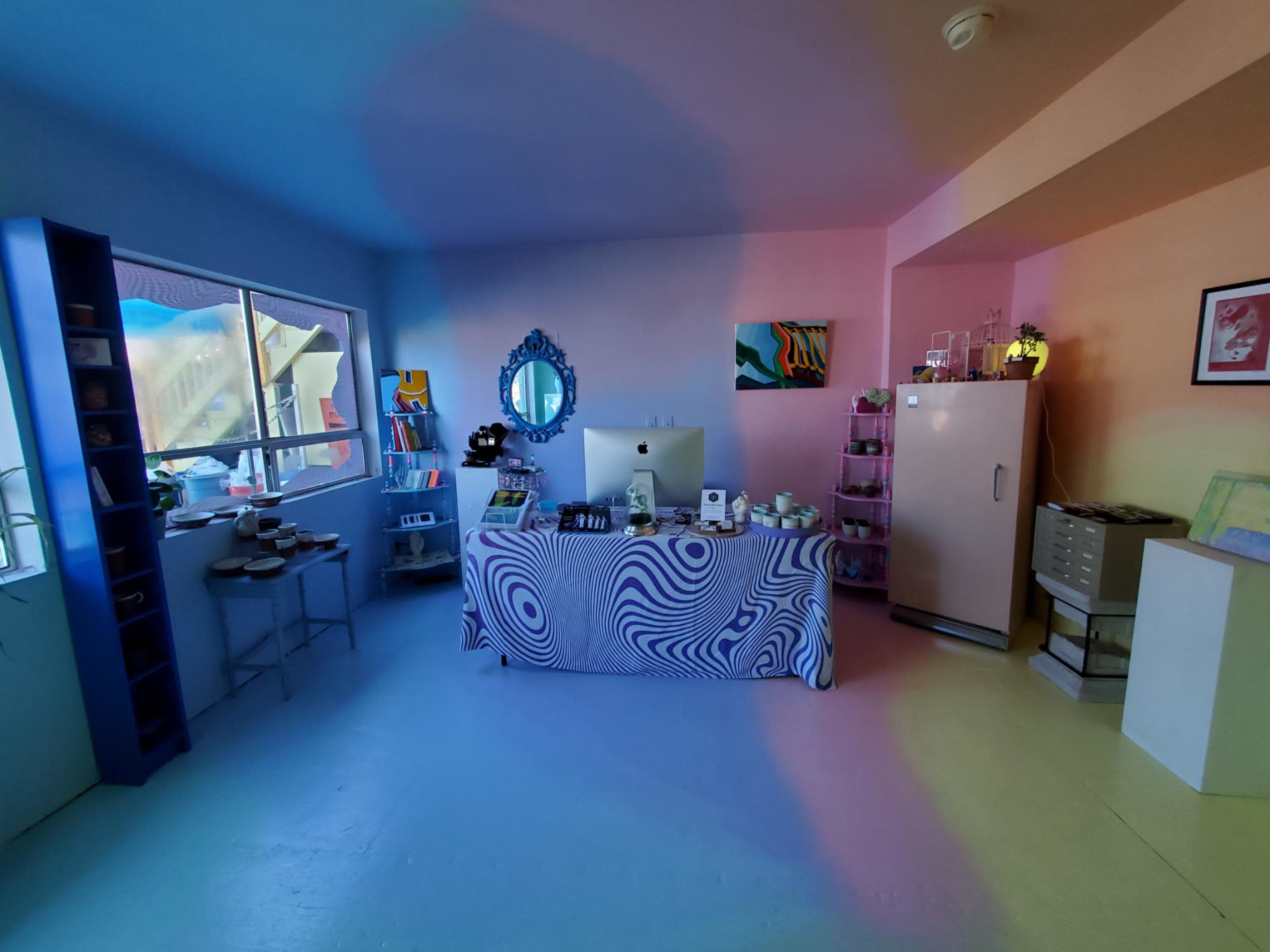 A colorful room features a patterned table at the center, a computer, a fridge, and various items displayed on shelves.