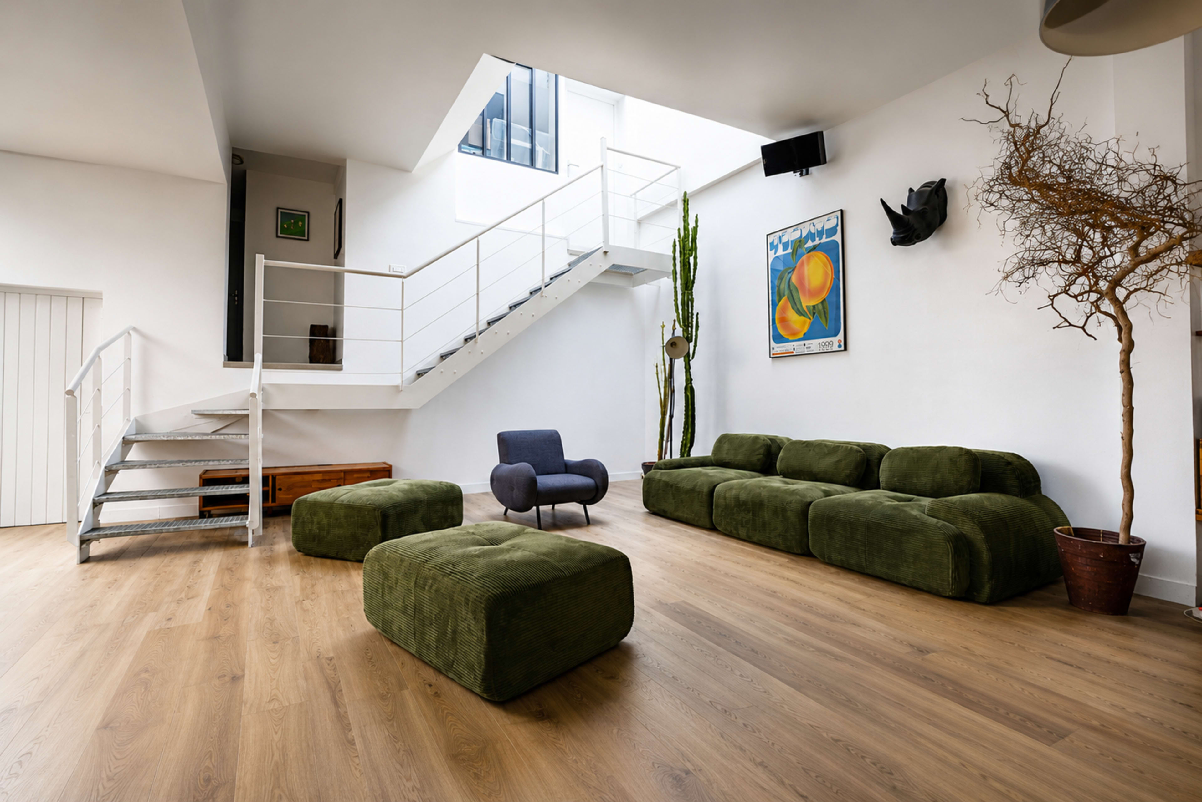 A bright, modern interior features a staircase, green upholstered seating, and a large window, highlighting minimalist decor and wooden flooring.