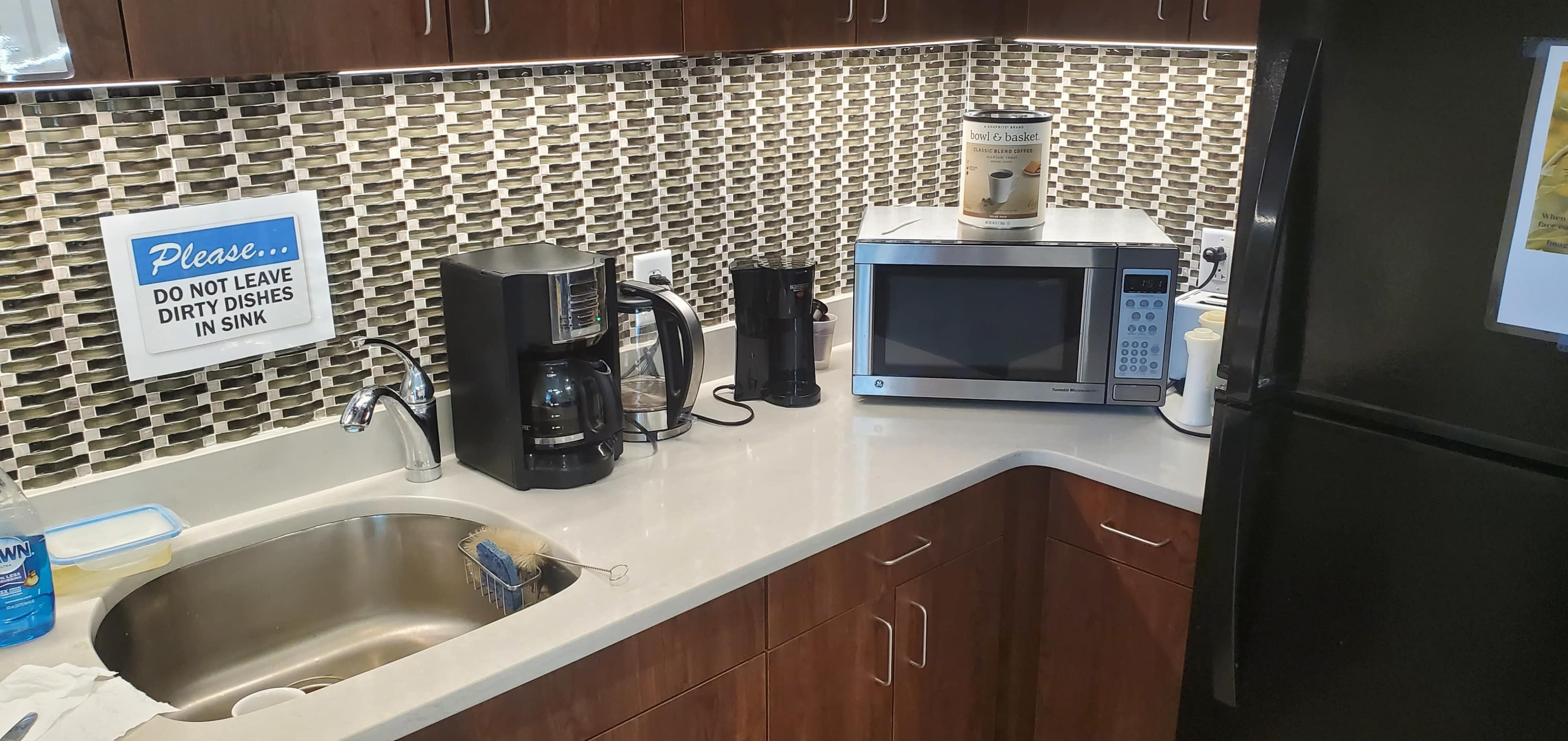 A kitchen countertop with a sink, coffee maker, microwave, and a canister on top, against a tiled backsplash.