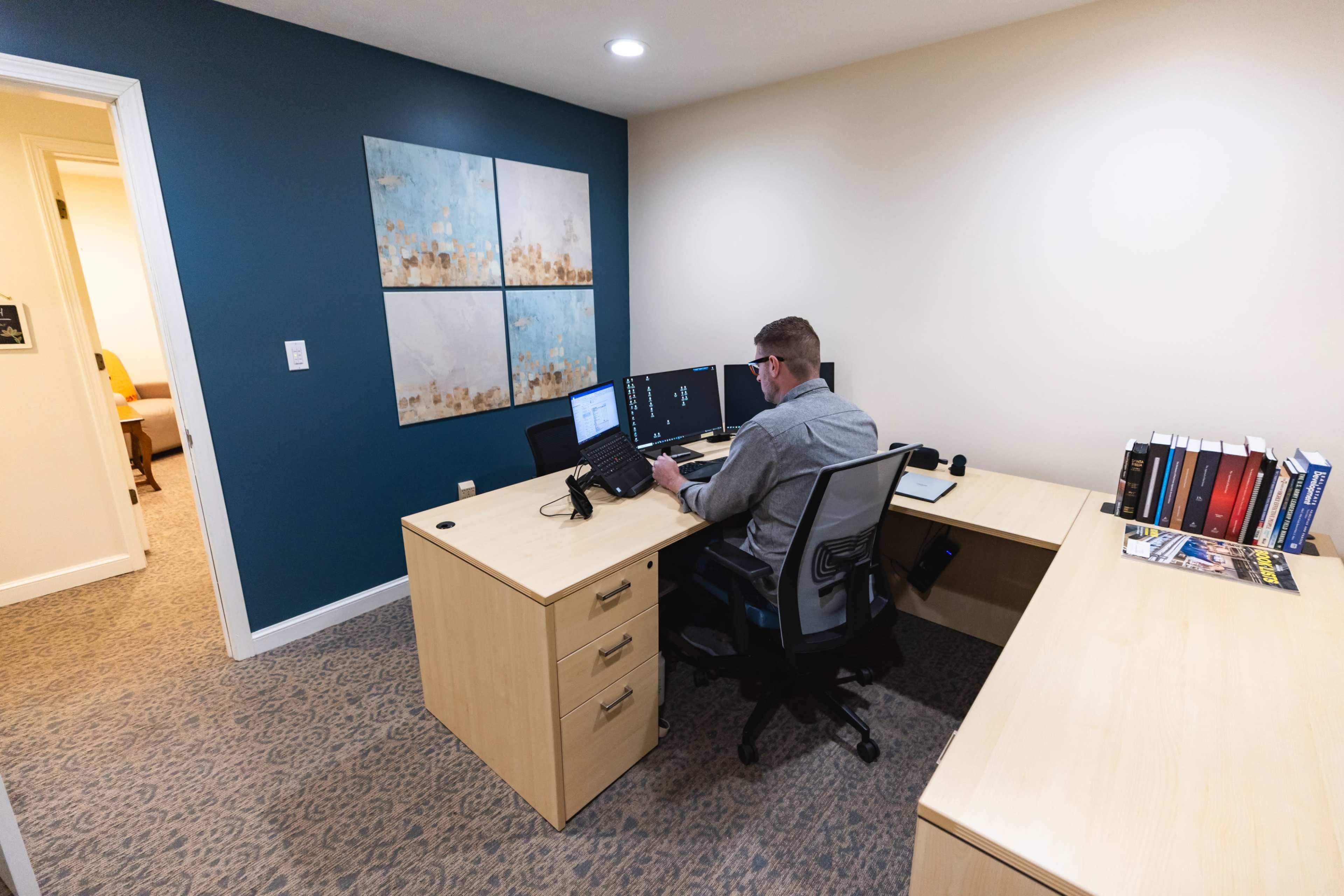 A person sits at a desk in an office, working on dual computer monitors, with bookcases and a doorway leading to another room visible in the background.