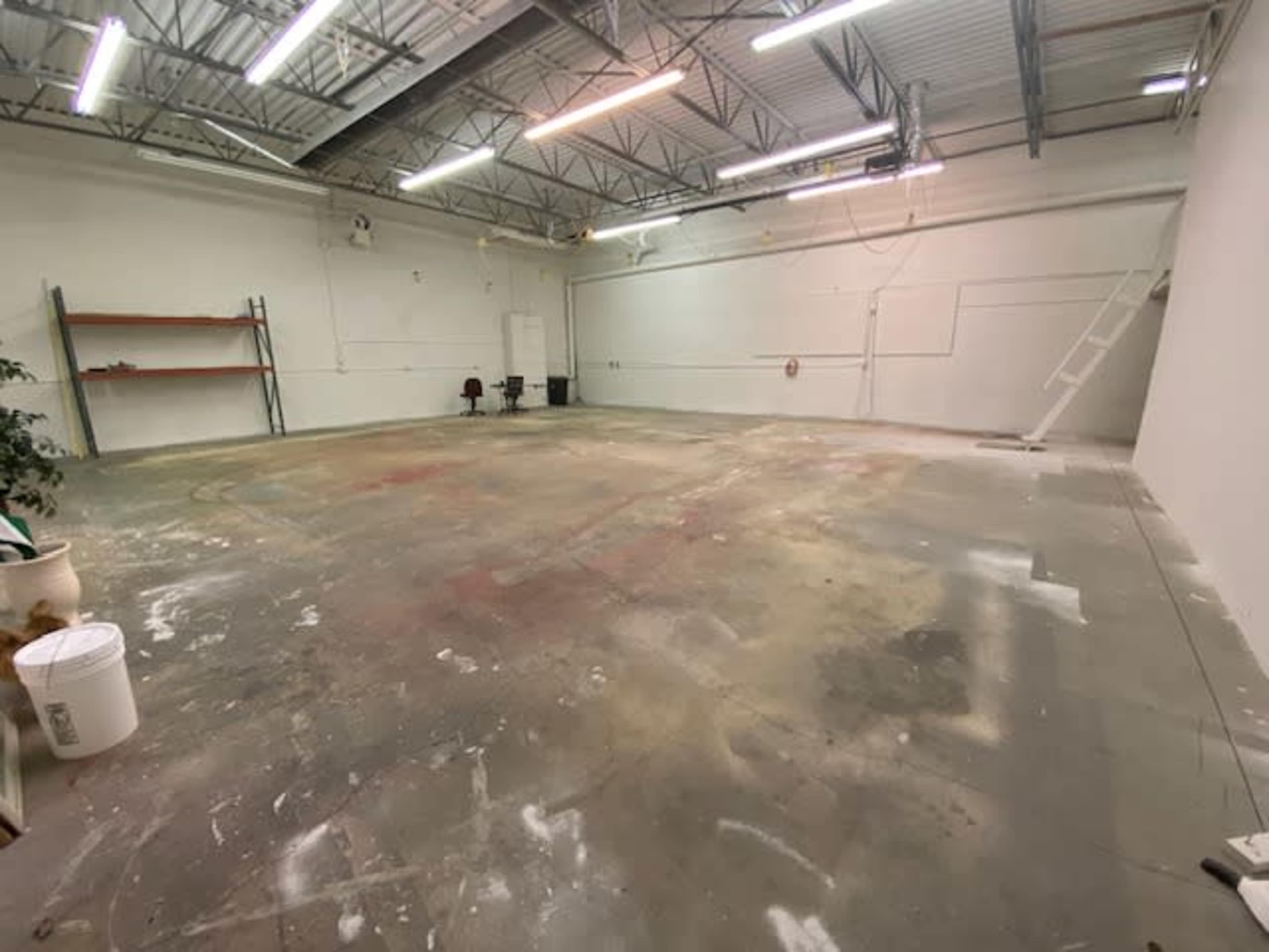 An empty industrial space with a concrete floor, metal shelving along one wall, and a staircase leading to a loft area.