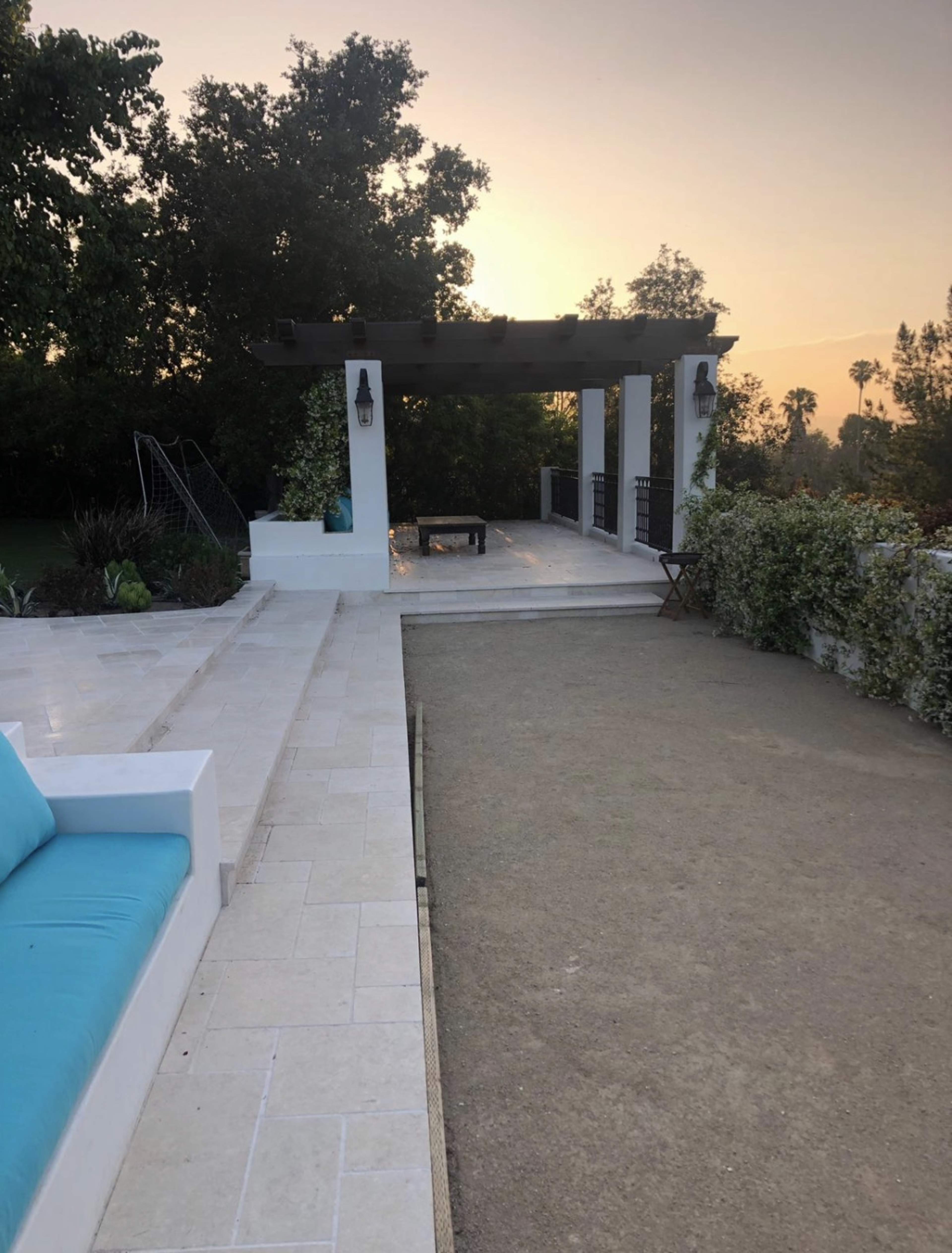 The image shows a patio area with a pergola, a stone walkway, and green shrubs under a sunset sky.