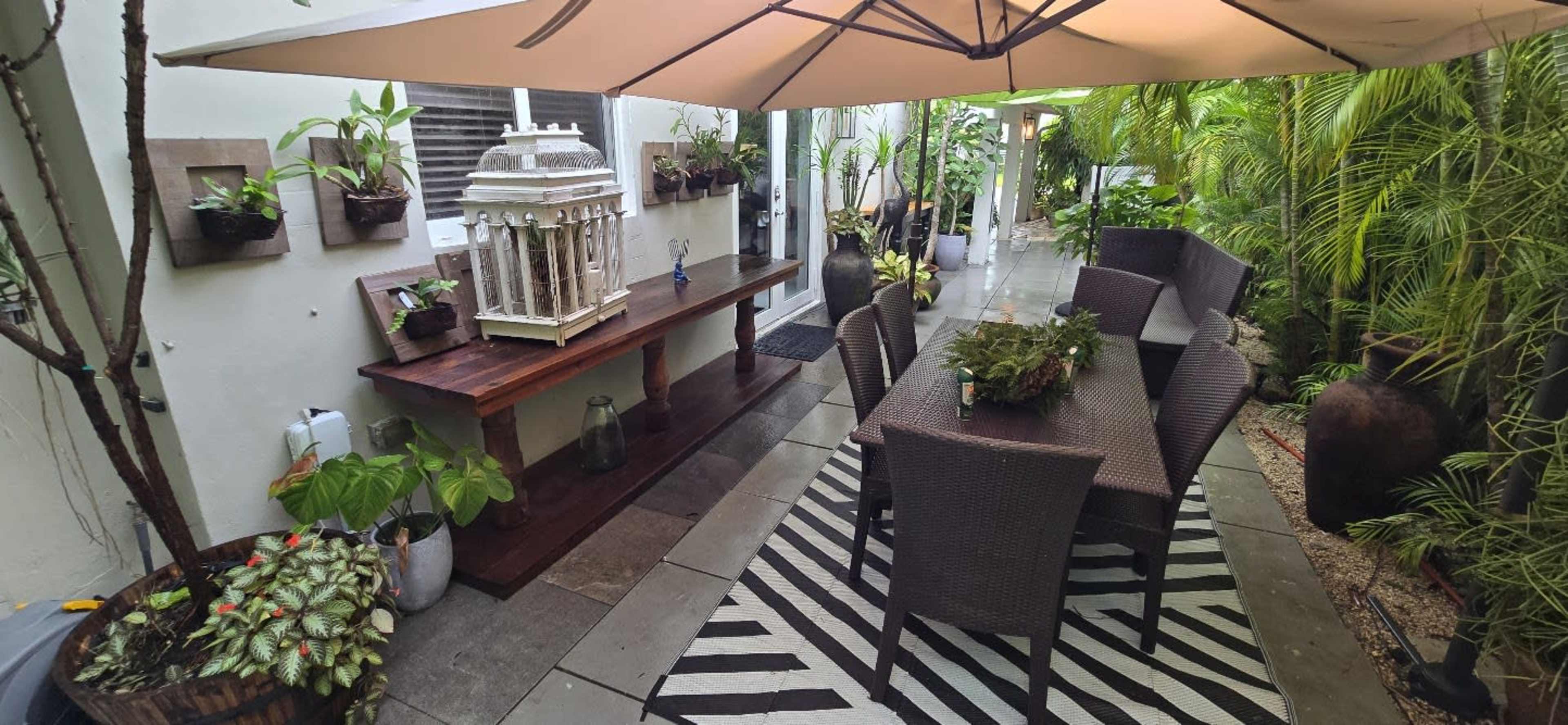The image shows a patio area featuring a large umbrella over a table surrounded by wicker chairs, with plants in pots and decorative elements along the walls.