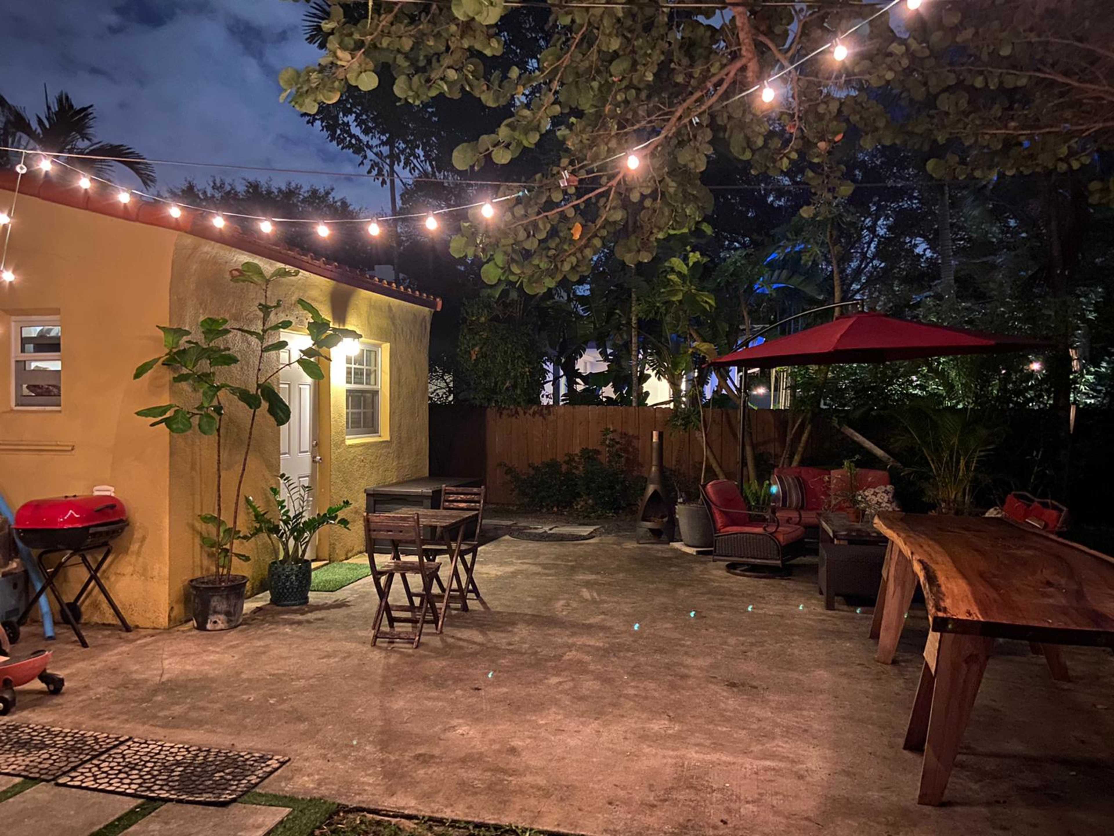 A backyard patio area featuring a wooden table, chairs, a grill, and string lights illuminating the space at night.